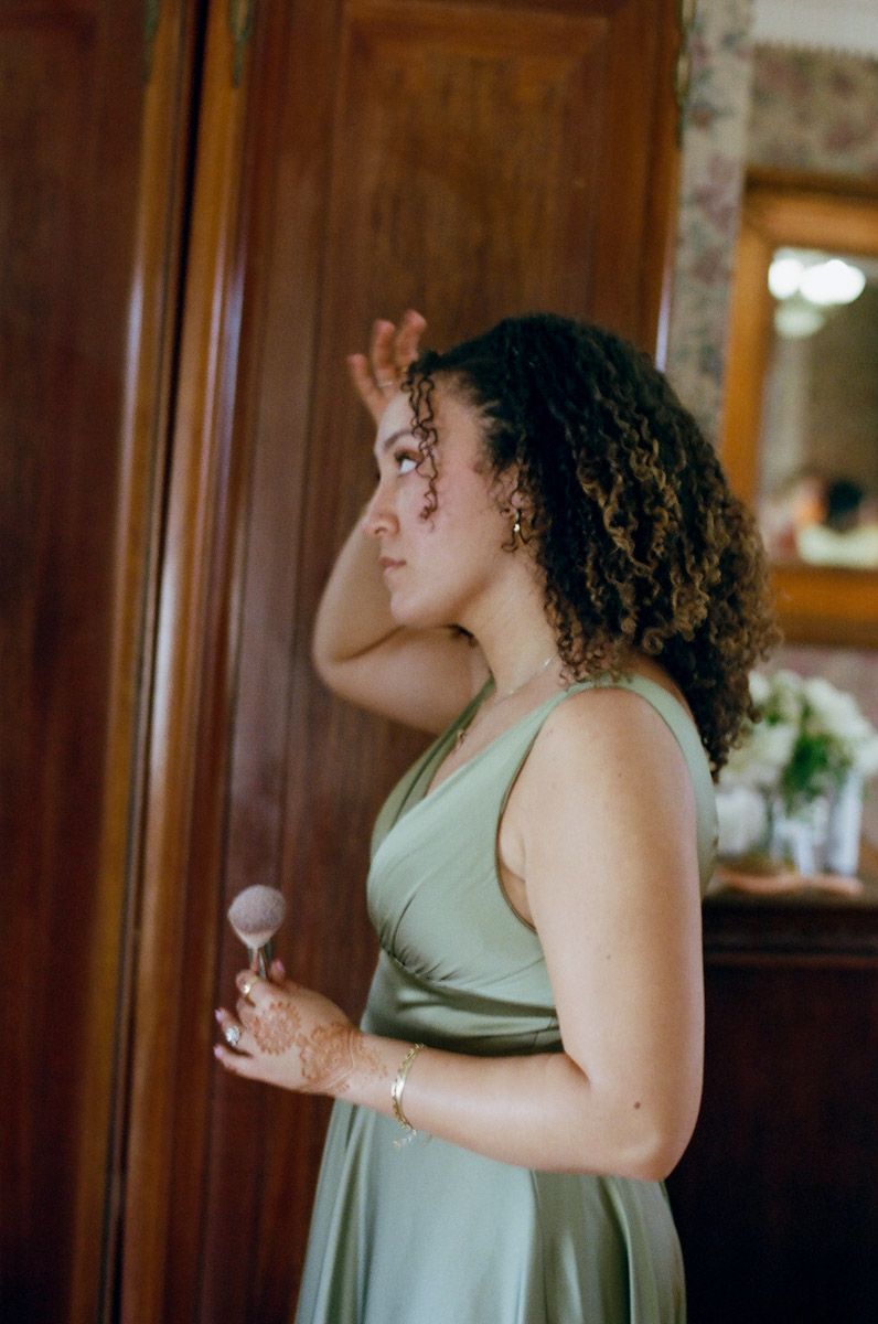 Bridesmaid in sage green dress holding a makeup brush while fixing hair in mirror.