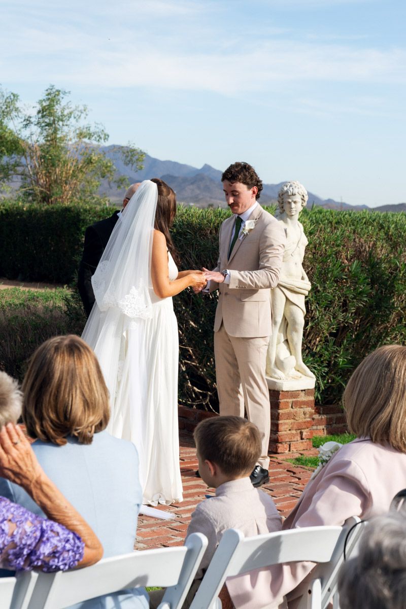 Outdoor wedding ceremony with bride in white gown and groom in tan suit exchanging rings before guests near a classical statue.