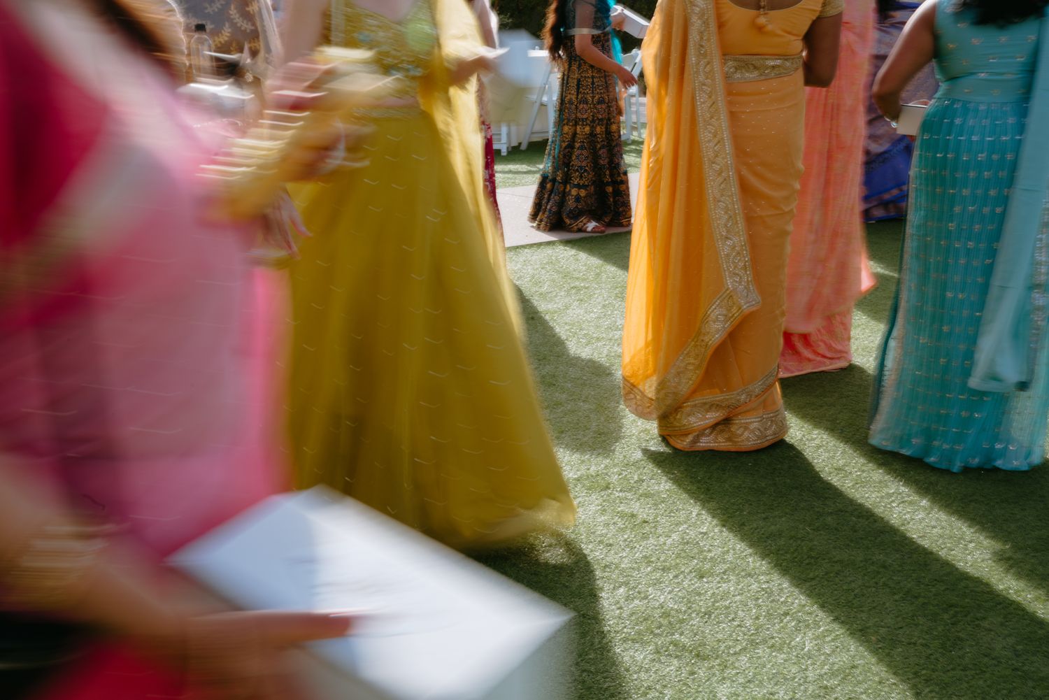 Blurred motion photo of women in colorful Indian lehengas and sarees standing on green grass at a wedding.