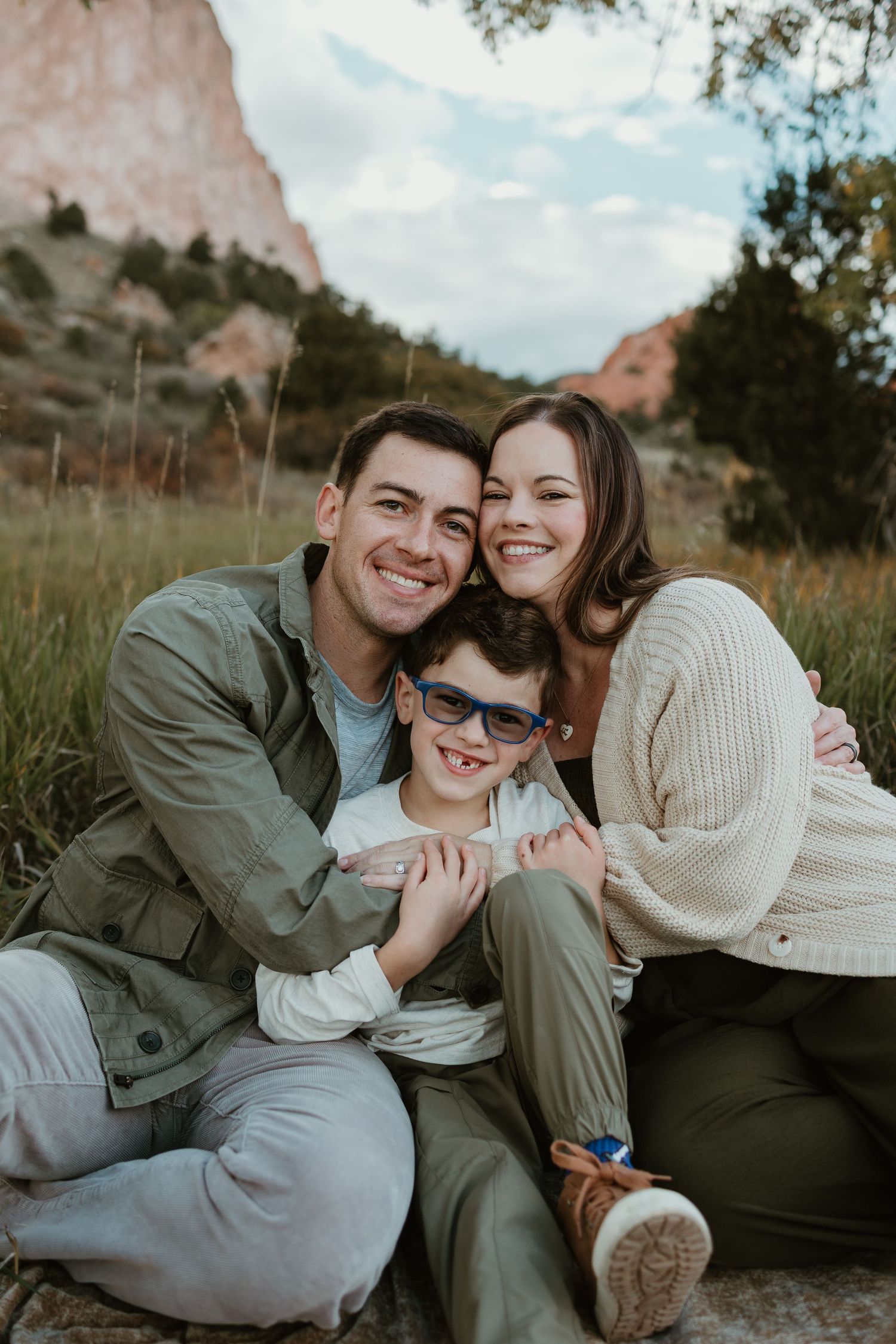 A smiling family of three poses outdoors near red rock formations, with parents embracing their young son wearing blue sunglasses.
