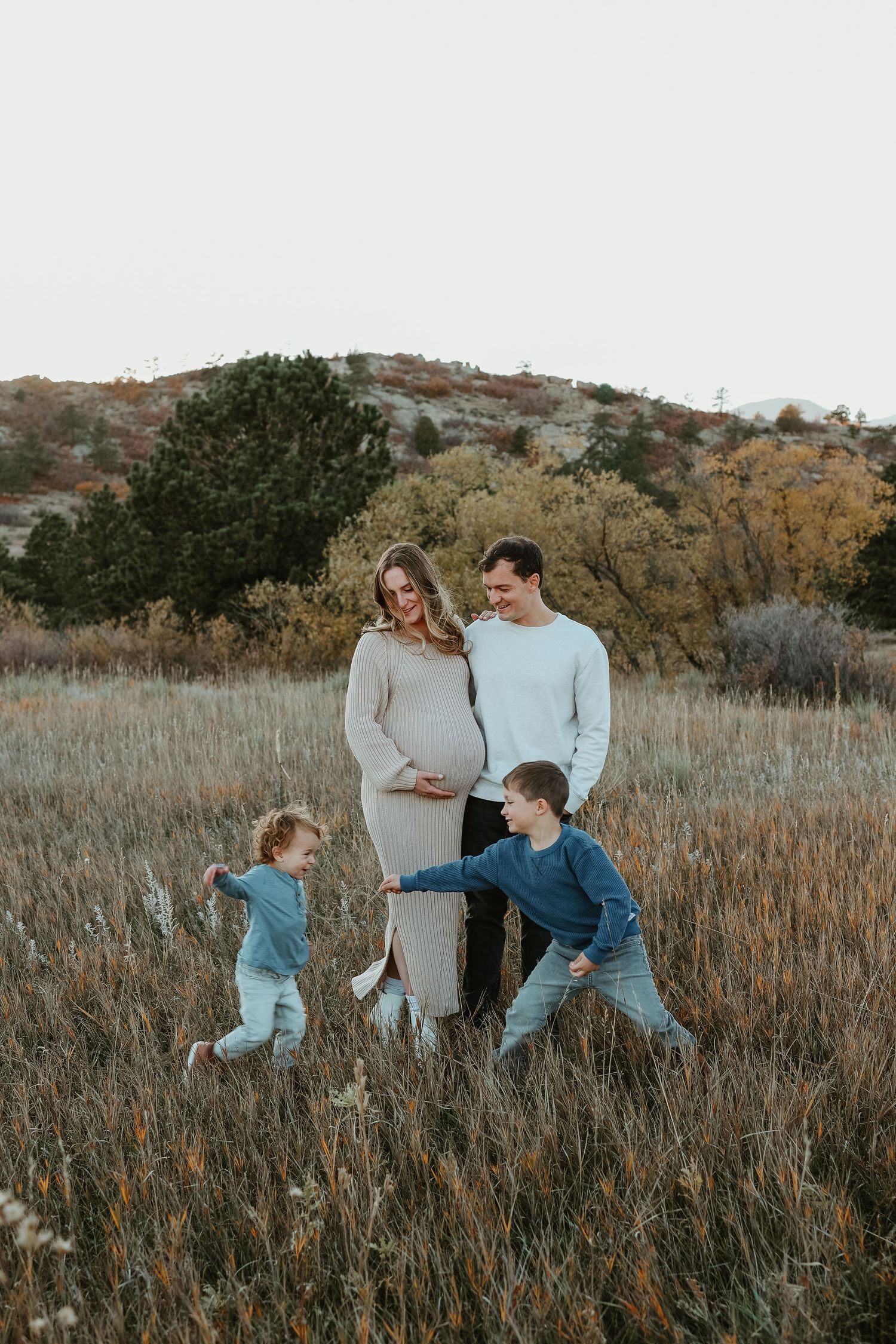 Pregnant woman and man stand in autumn field while two young children run toward them during maternity photo session.