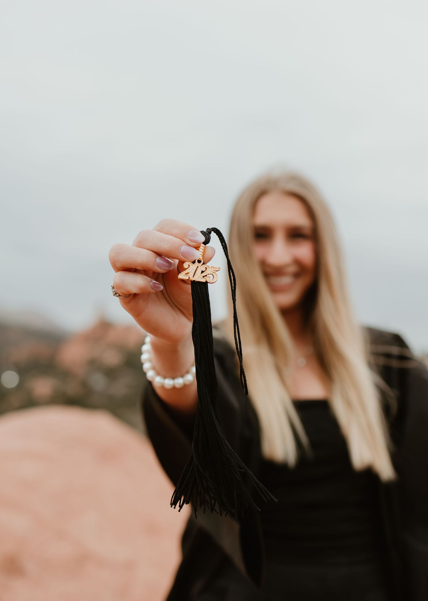 A hand wearing a pearl bracelet holds up a sparkling engagement ring against a blurred outdoor landscape background.