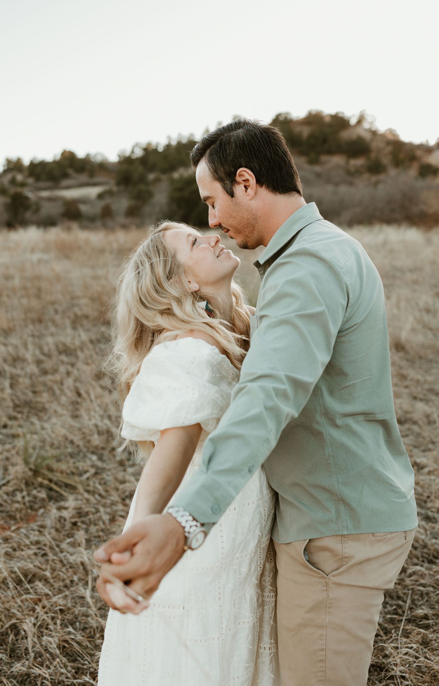 A romantic couple in a field shares an intimate moment at sunset, with the woman in white and man in light blue.