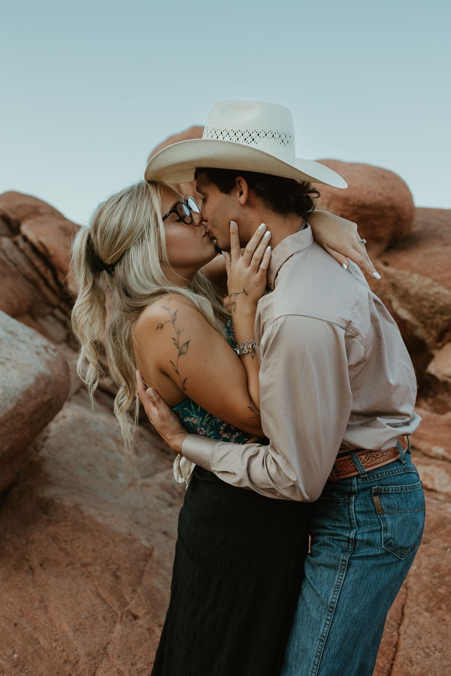 A couple sharing a romantic kiss in a desert landscape, the man wearing a white cowboy hat and the woman with blonde hair and tattoos.