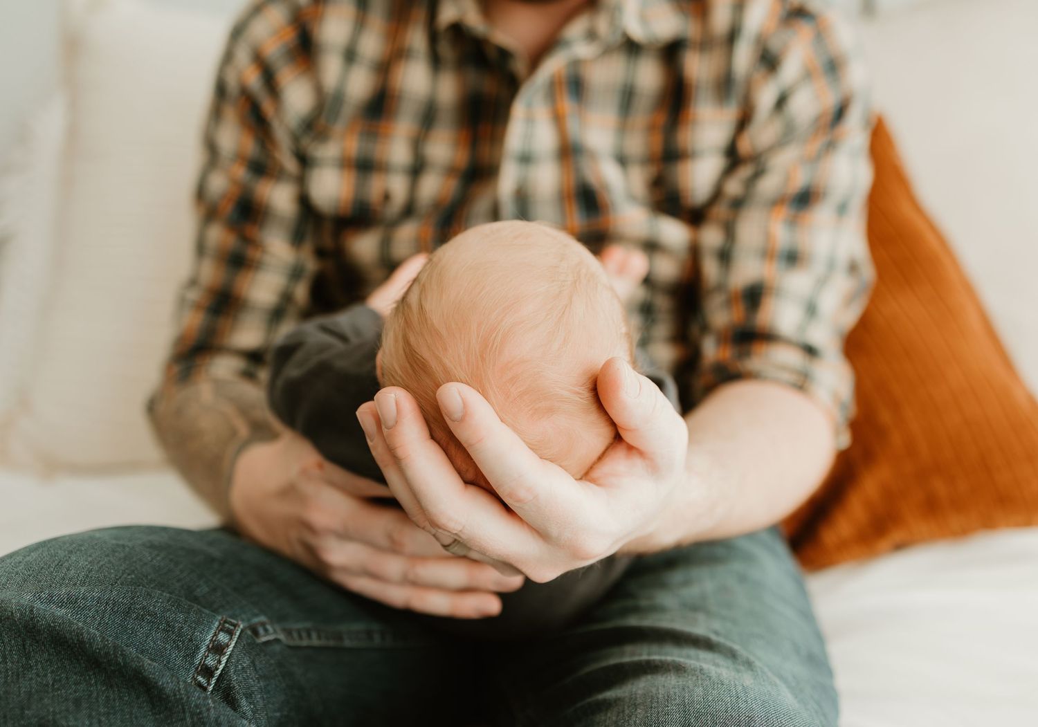 Someone sitting on a couch wearing plaid shirt and jeans holds a sleeping newborn baby.