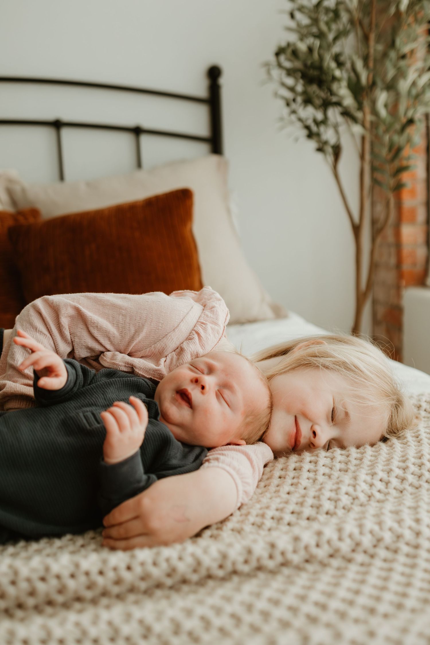 Two small children sleeping peacefully together on a cozy knitted blanket with a rustic headboard and plant decor in background.