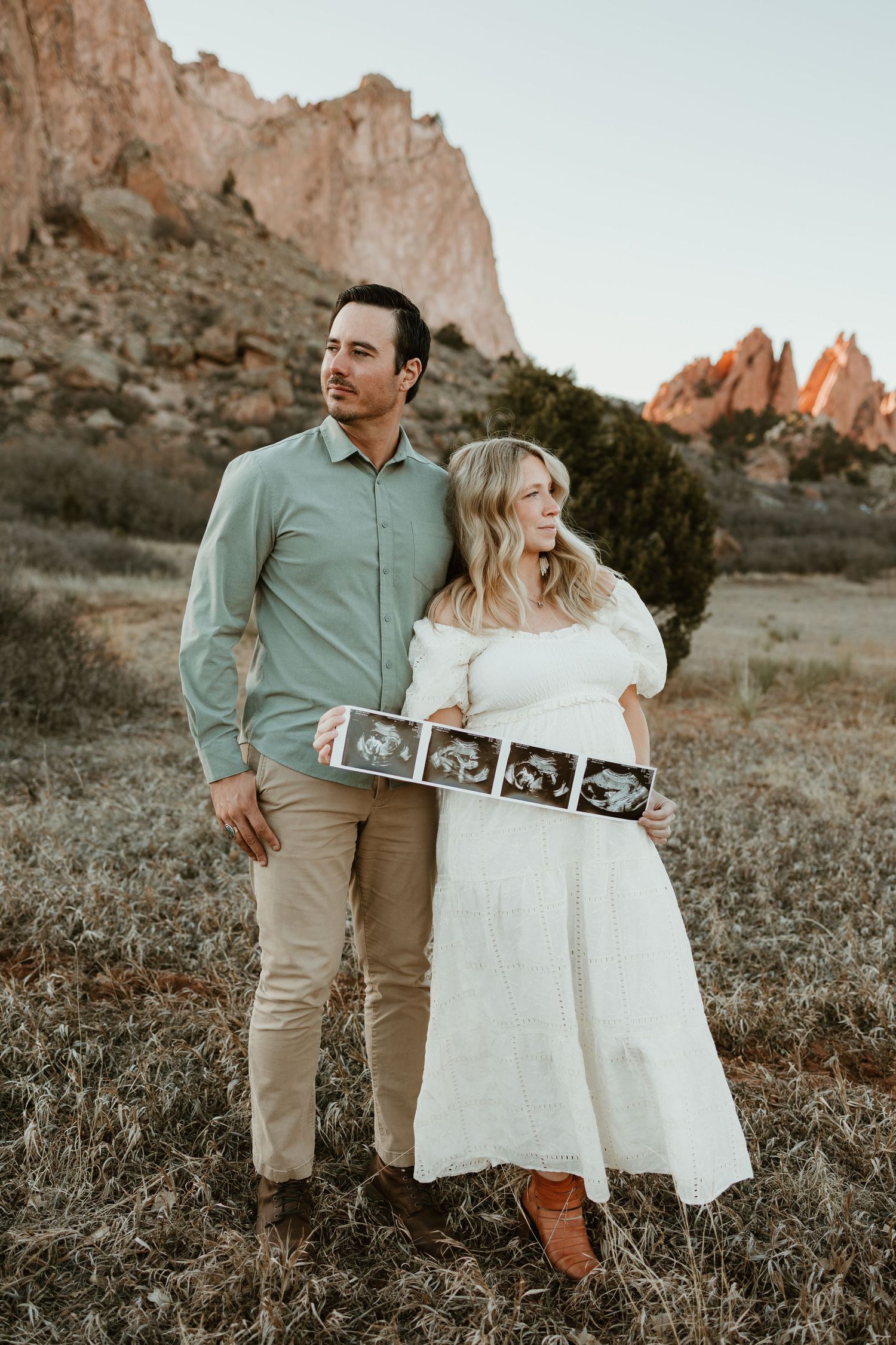 A couple stands in front of rocky mountains holding ultrasound photos during sunset at Garden of the Gods.
