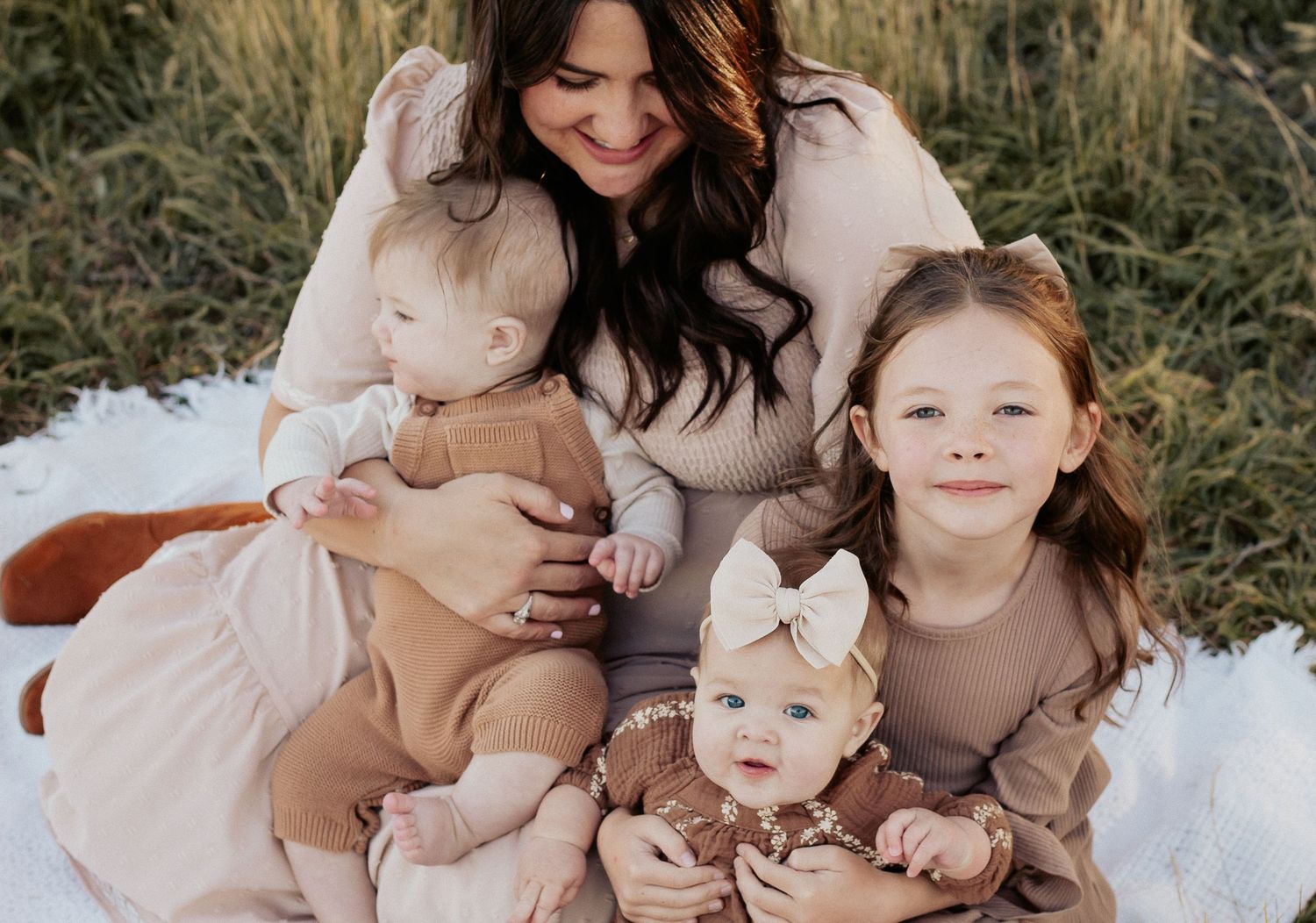 Candid family portrait with mother and children sitting on blanket in field during golden hour.