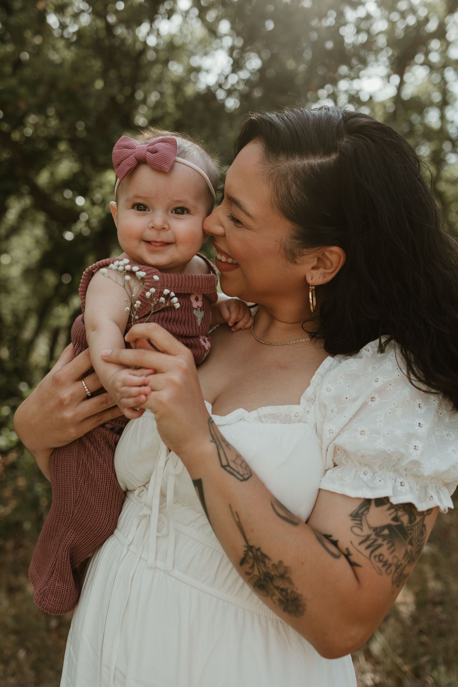 A tattooed mother in a white dress lovingly holds her smiling baby girl wearing a pink bow outdoors.