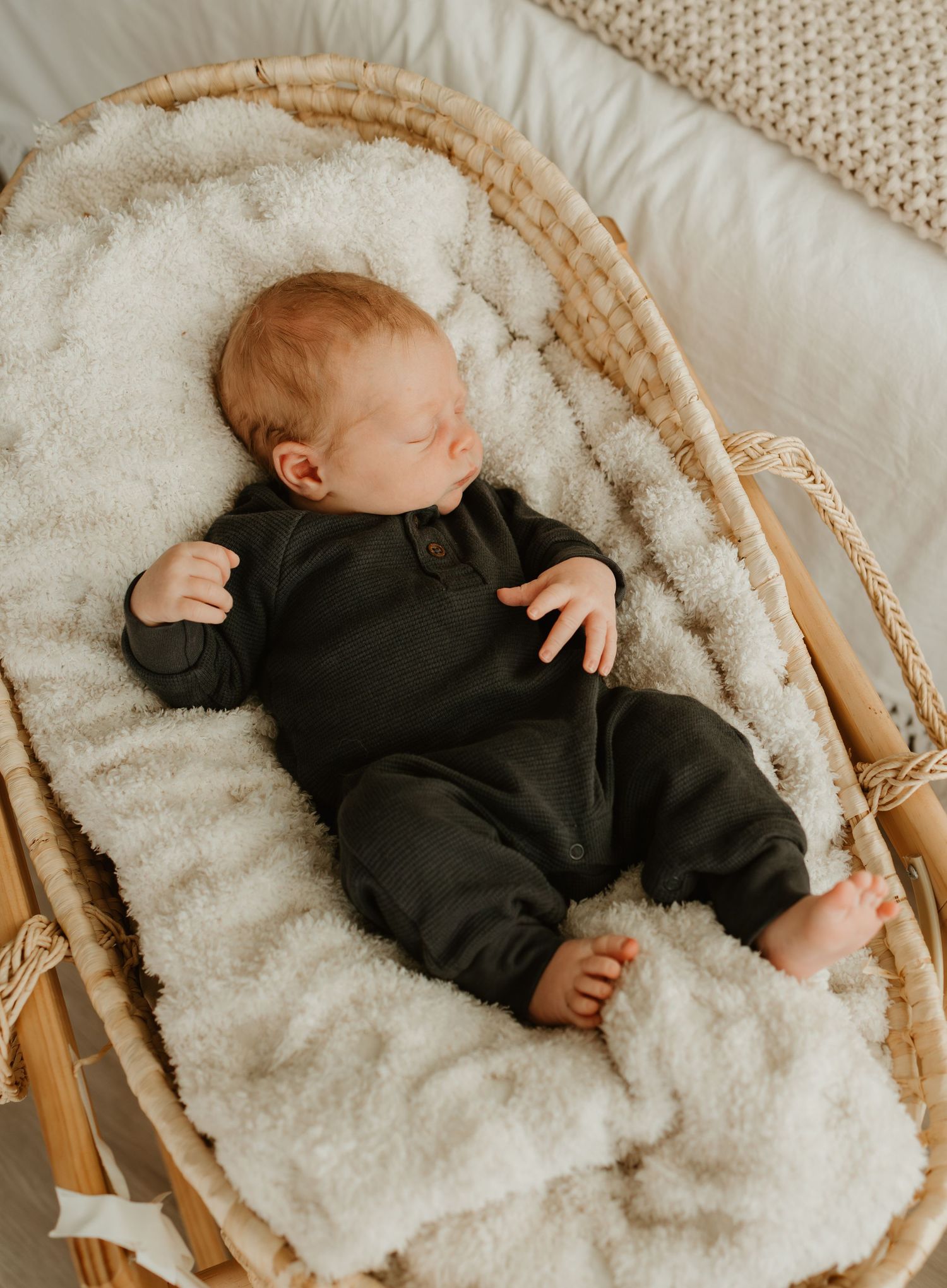 Baby sleeping peacefully in a cozy wicker bassinet lined with plush white sheepskin and wearing a black outfit.
