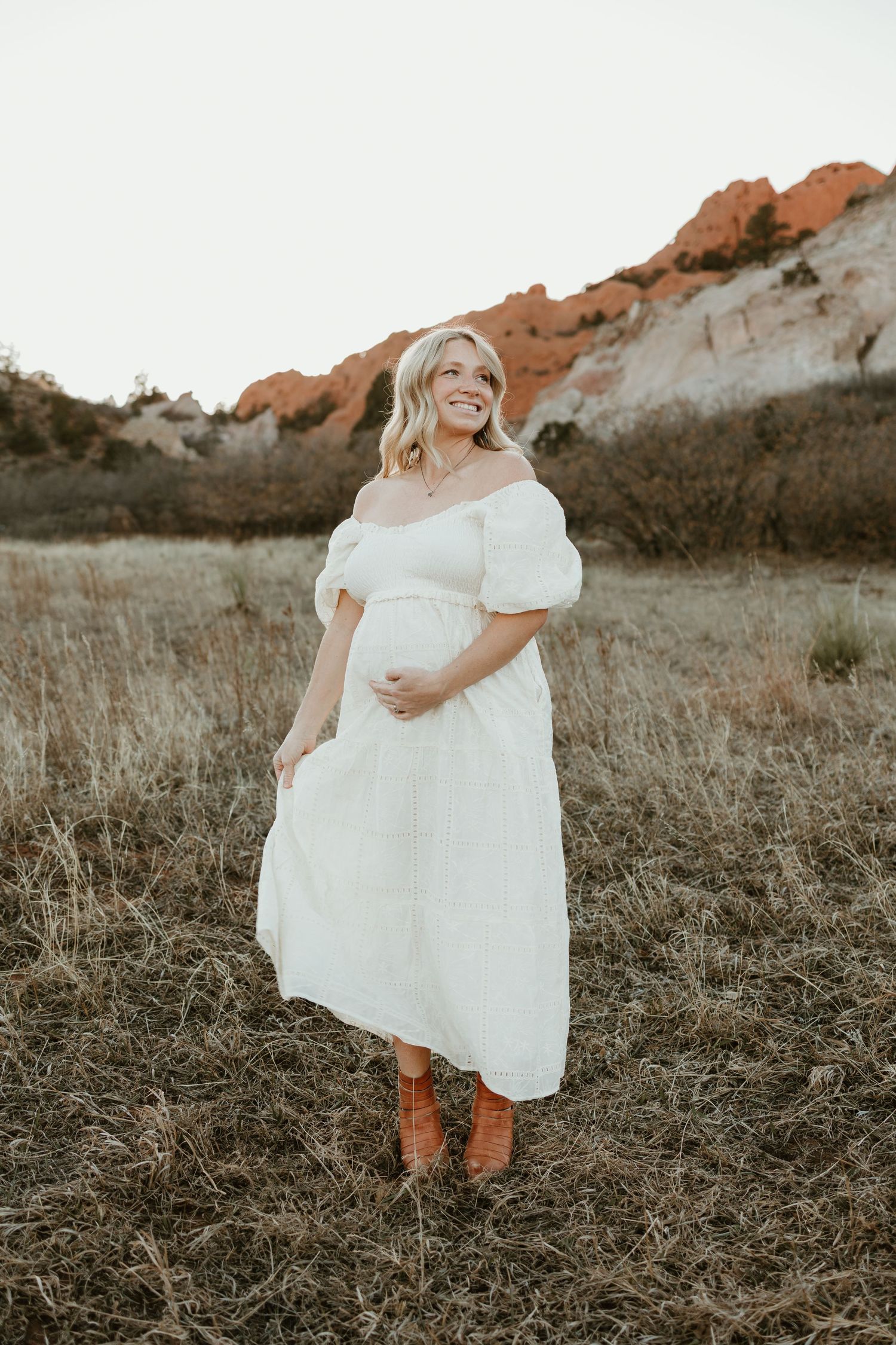 A woman in a white off-shoulder dress stands in a field with red rock formations visible in the background at sunset.