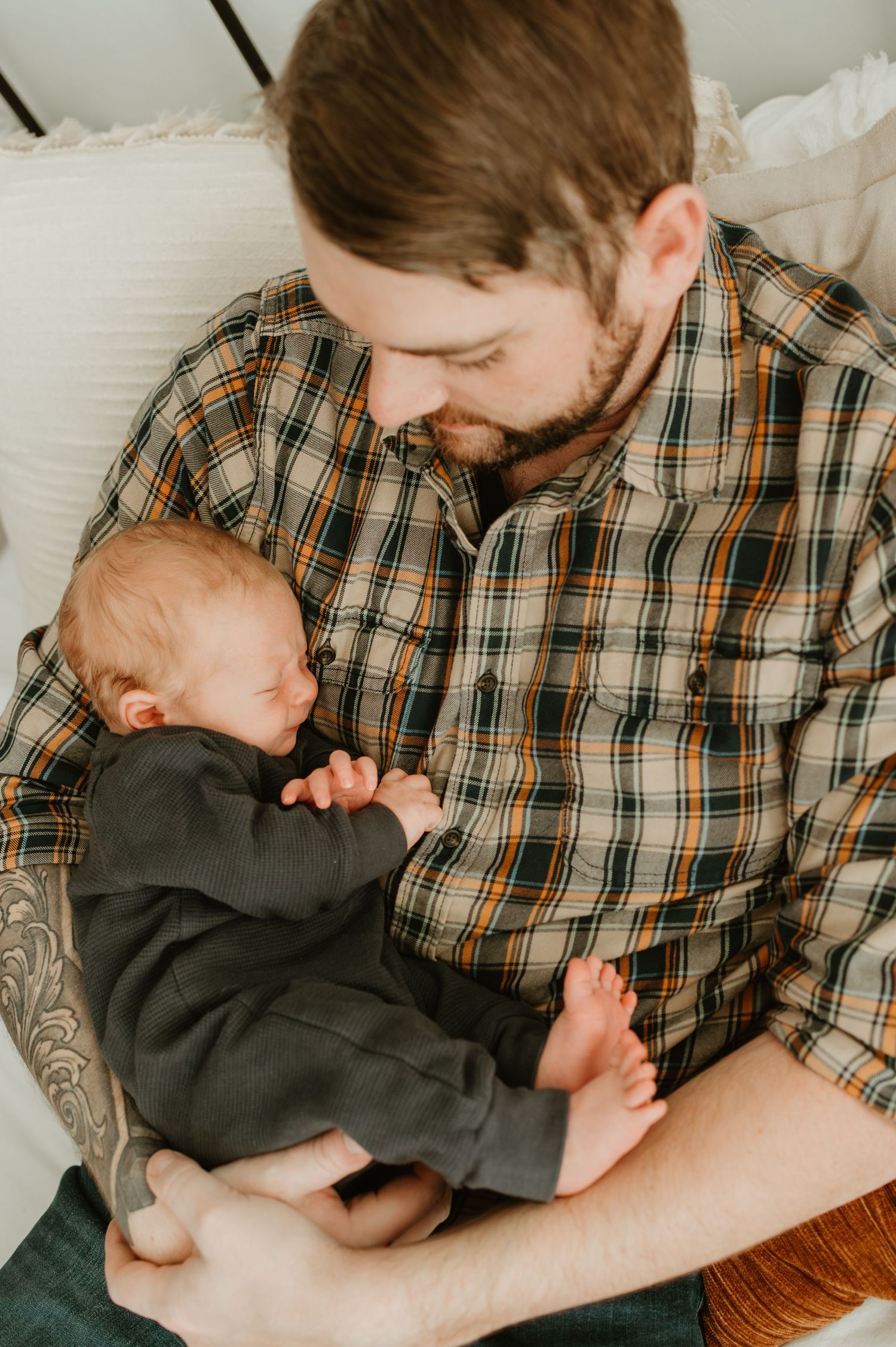 A tender moment between parent in plaid shirt and sleeping infant in dark clothing resting on upholstered chair.