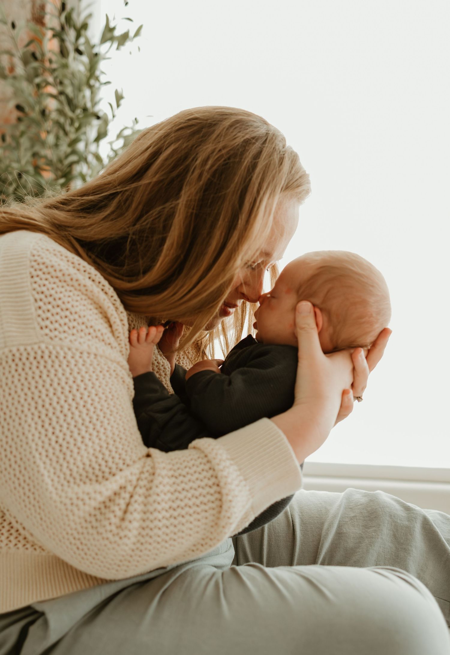 A person in a cream sweater holds a baby while sitting by a window in warm natural lighting.