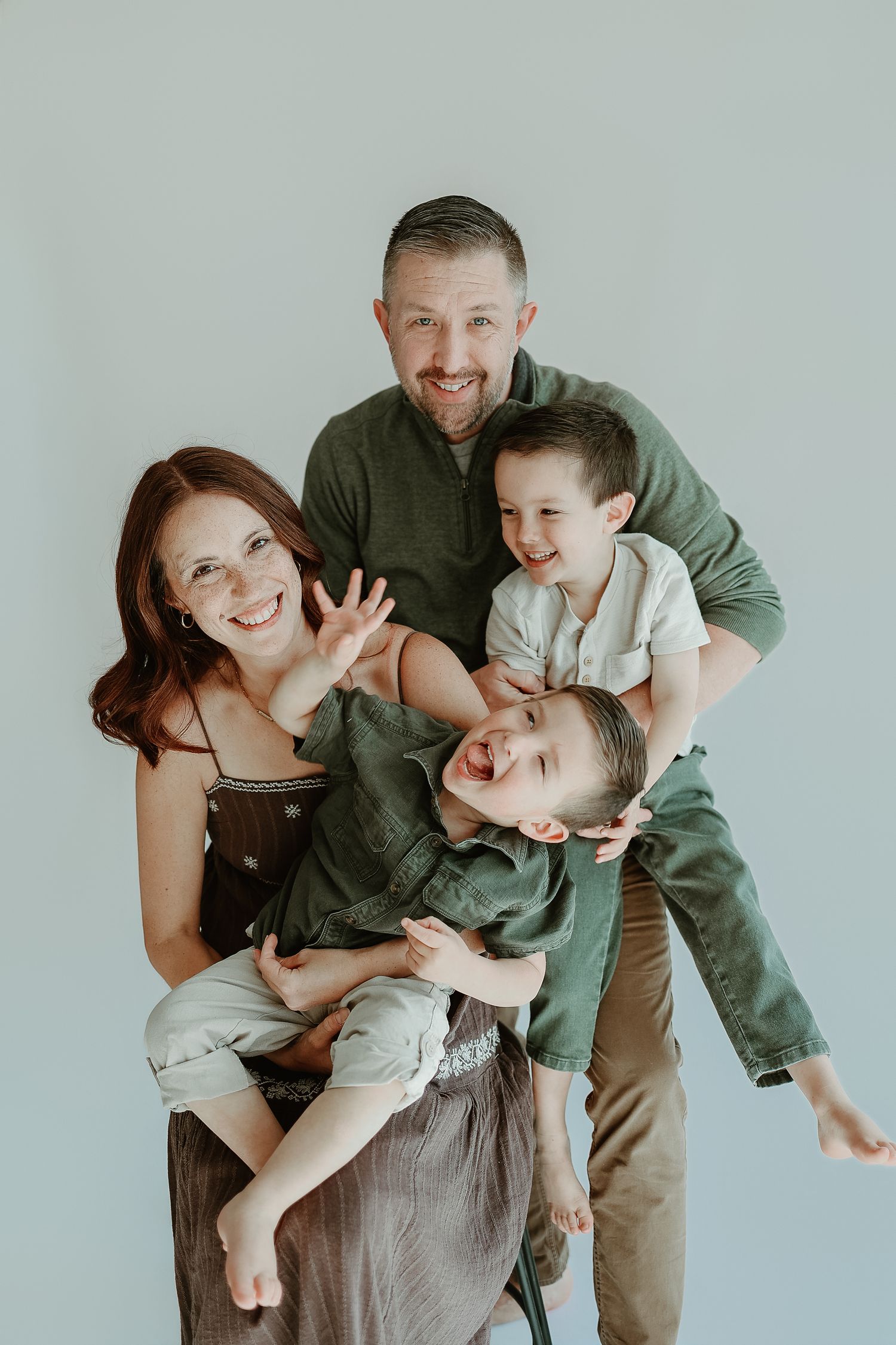 A joyful family of four poses together, with parents holding two laughing young boys against a light gray background.