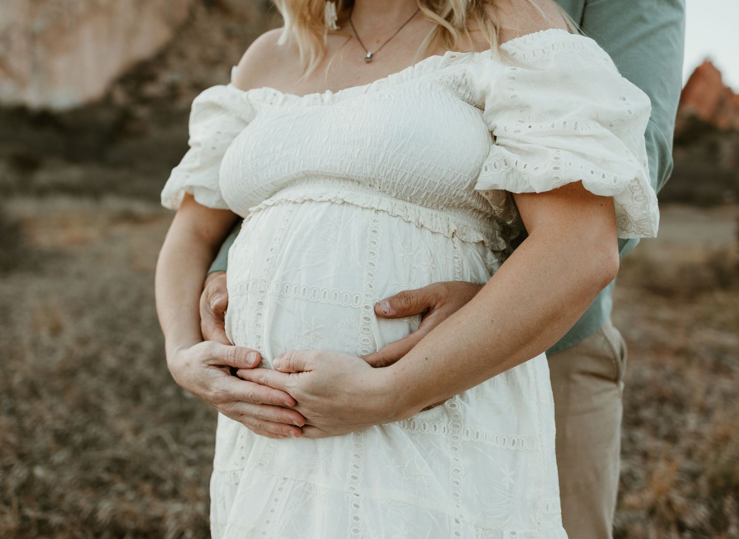 A pregnant woman in a flowing white dress tenderly holds her belly during a sunset maternity photoshoot outdoors.