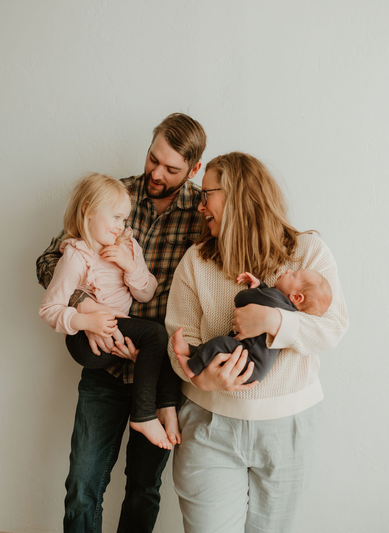 A family in casual clothing stands together against a light gray wall in a minimalist indoor portrait setting.