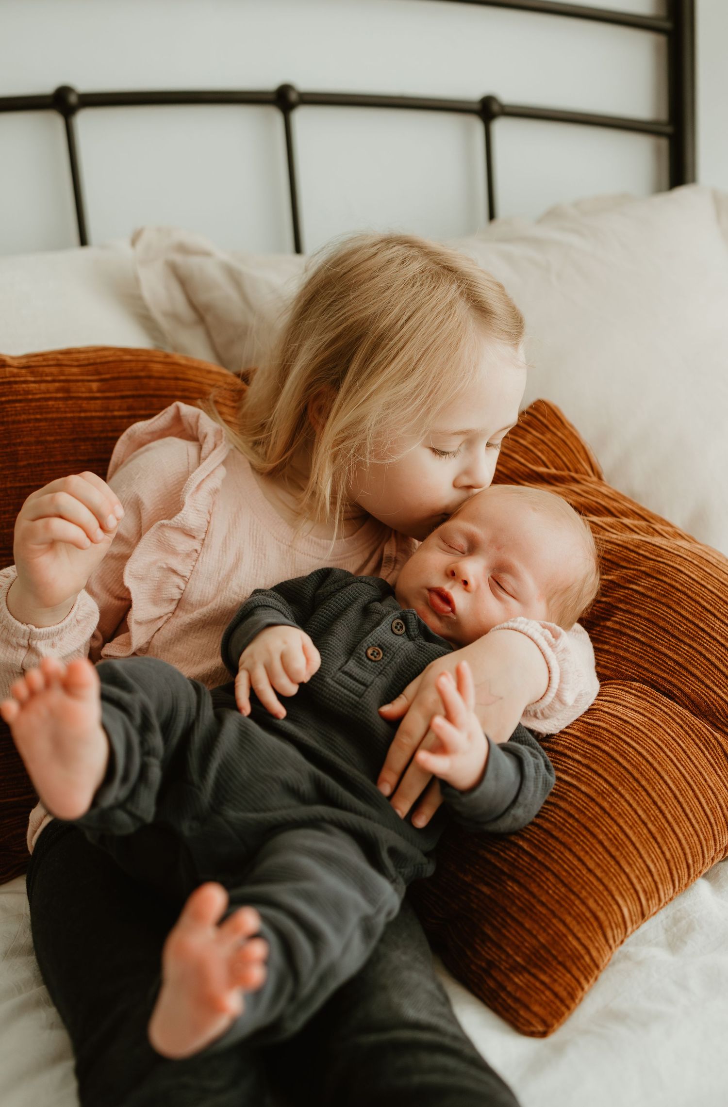 Siblings share tender moment on orange velvet pillow as toddler gently embraces baby in dark outfit.