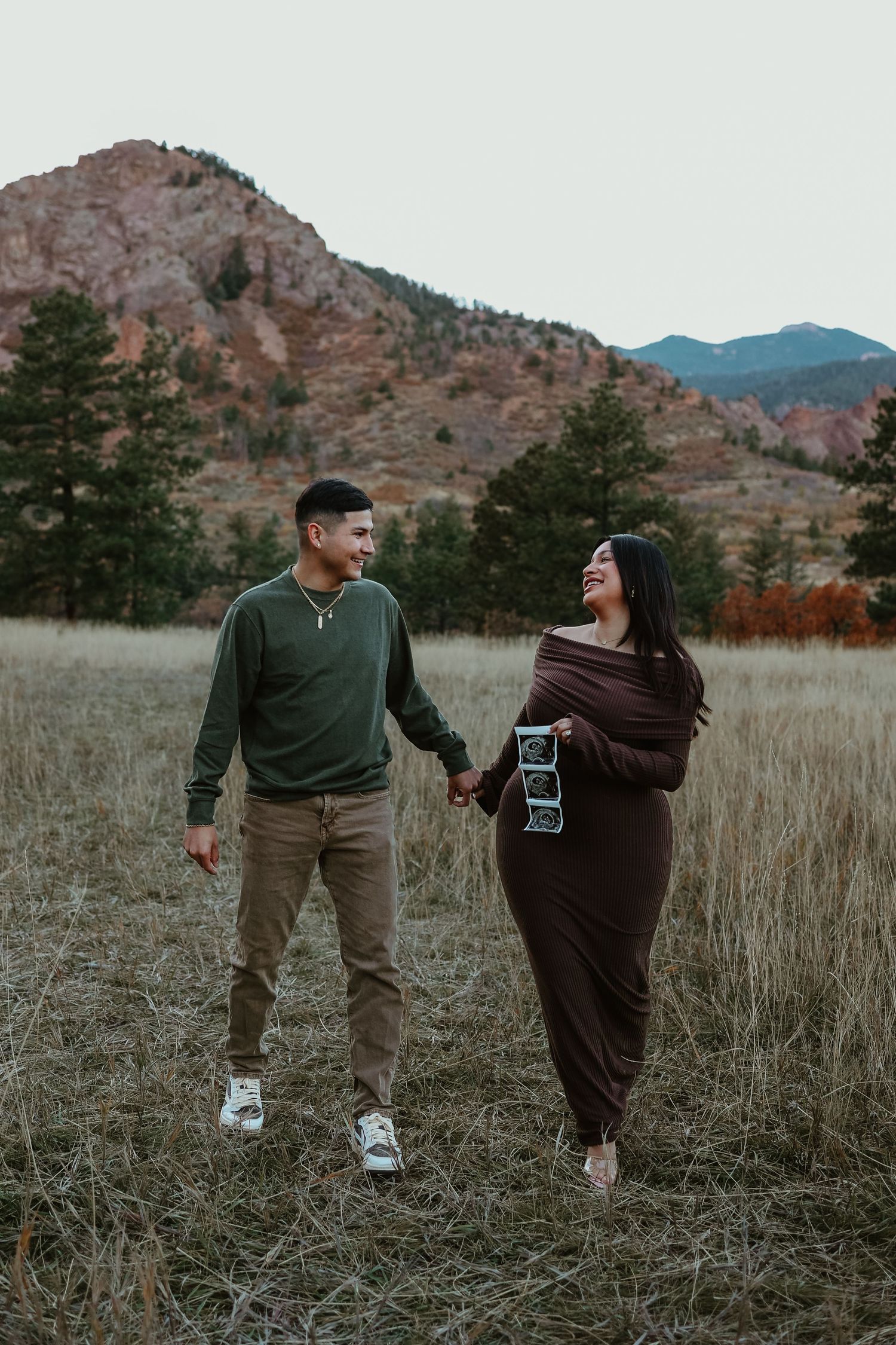 Couple holding hands in a mountain meadow, woman in brown maternity dress holding ultrasound photos, fall scenery.