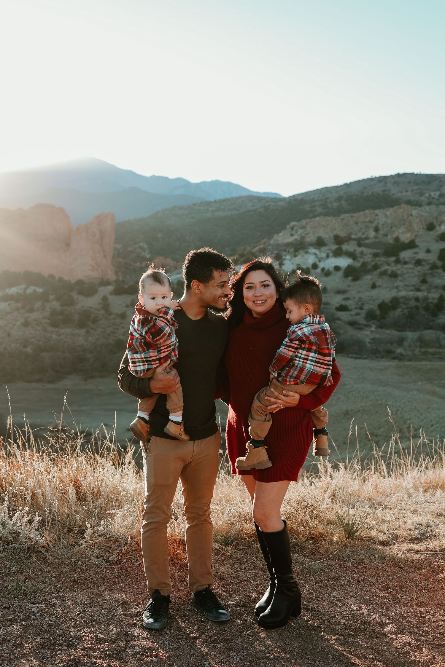 Family of four posing outdoors at golden hour with mountain landscape, parents holding two young children in plaid shirts.