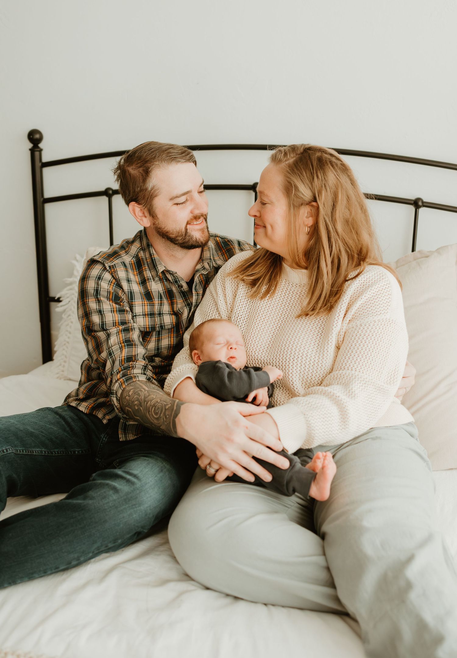 A couple sits together on a bed with their newborn baby, sharing an intimate family moment in soft natural light.