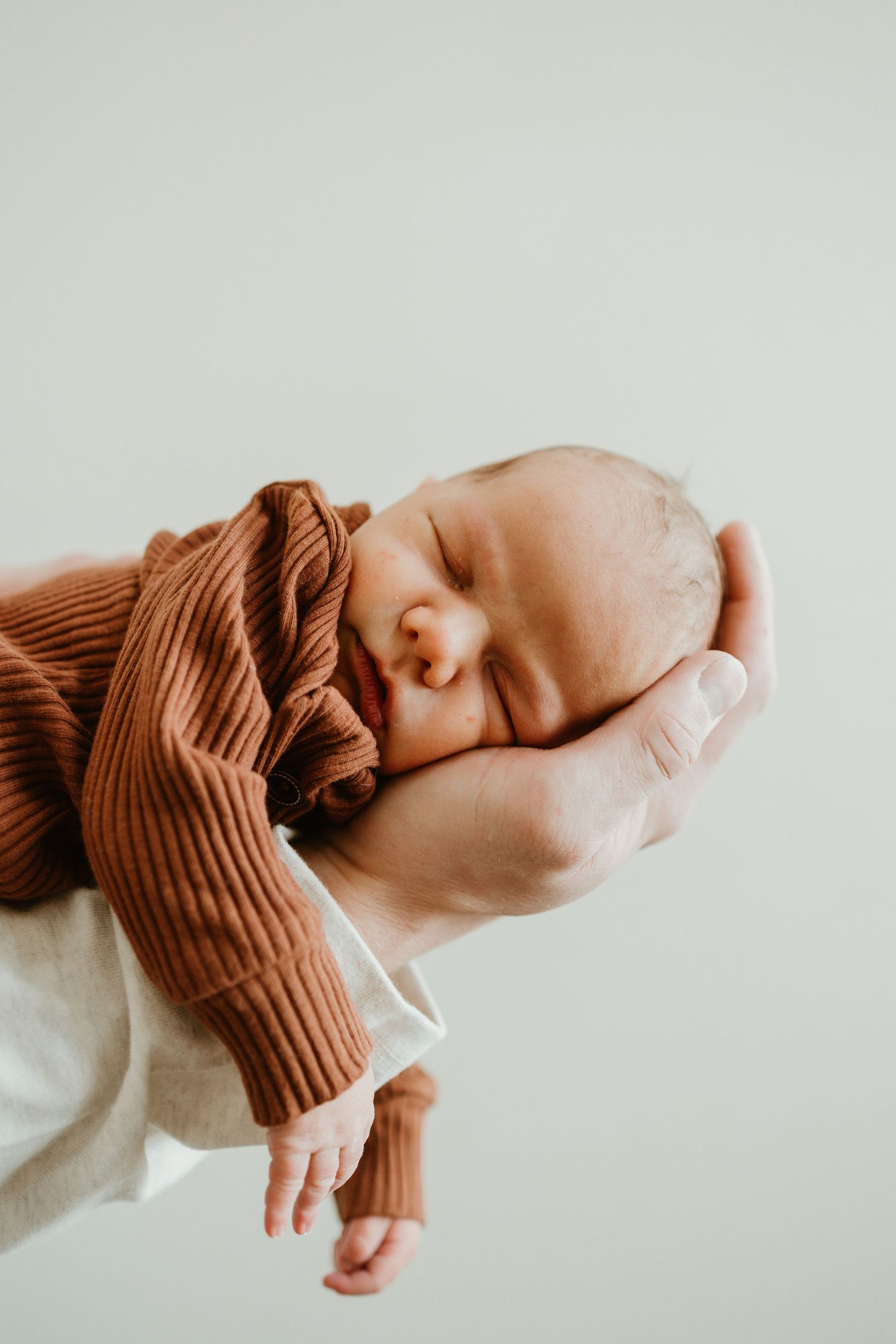 A peaceful sleeping newborn wrapped in a brown knit sweater rests gently in caring hands against a white background.