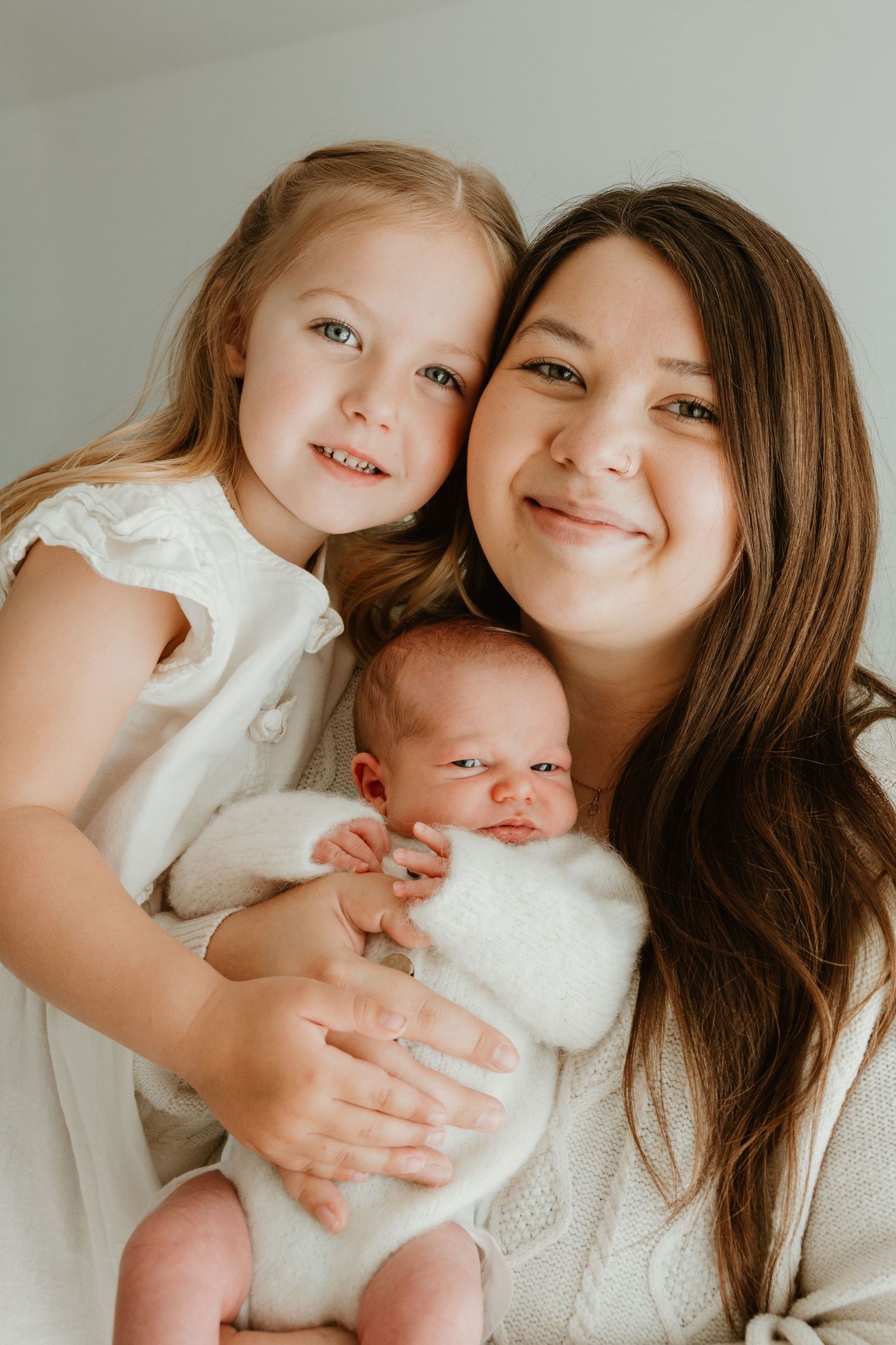 A smiling mother holds her newborn baby wrapped in white while her young blonde daughter poses beside them.