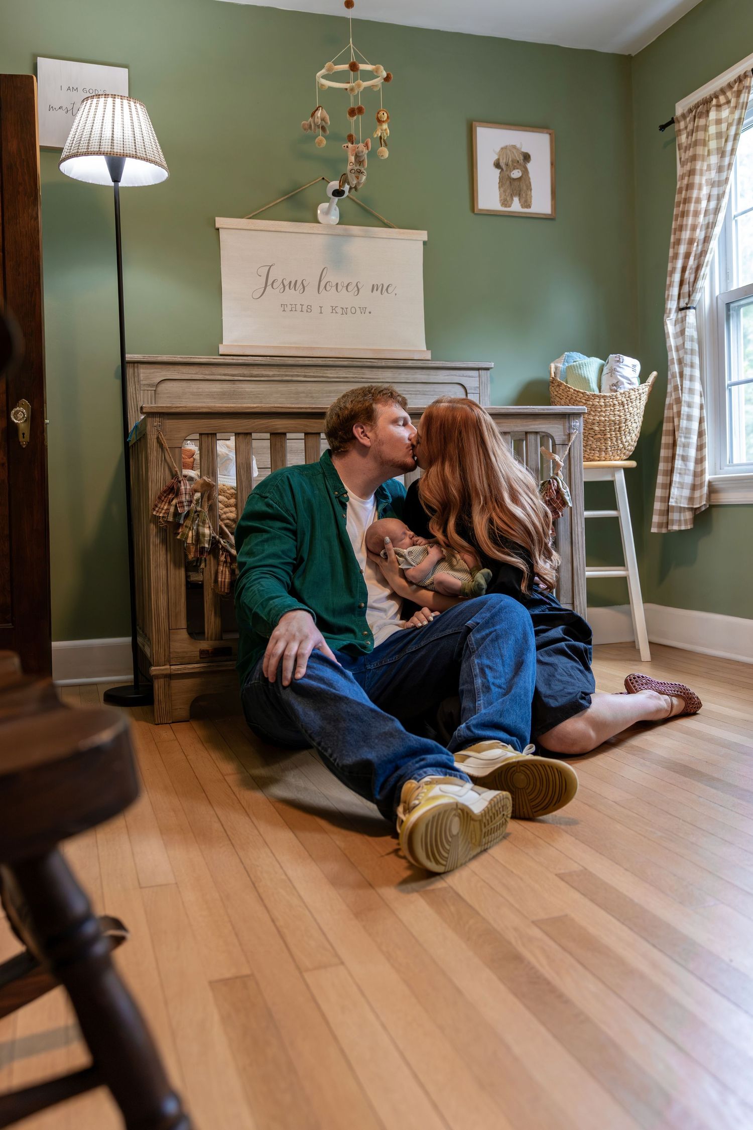 Couple sitting on hardwood floor kissing in a cozy green nursery with a crib and Jesus loves me sign.