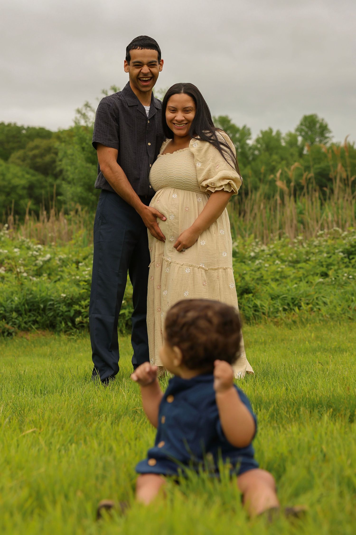 Smiling pregnant couple stands in green field while toddler sits in foreground.