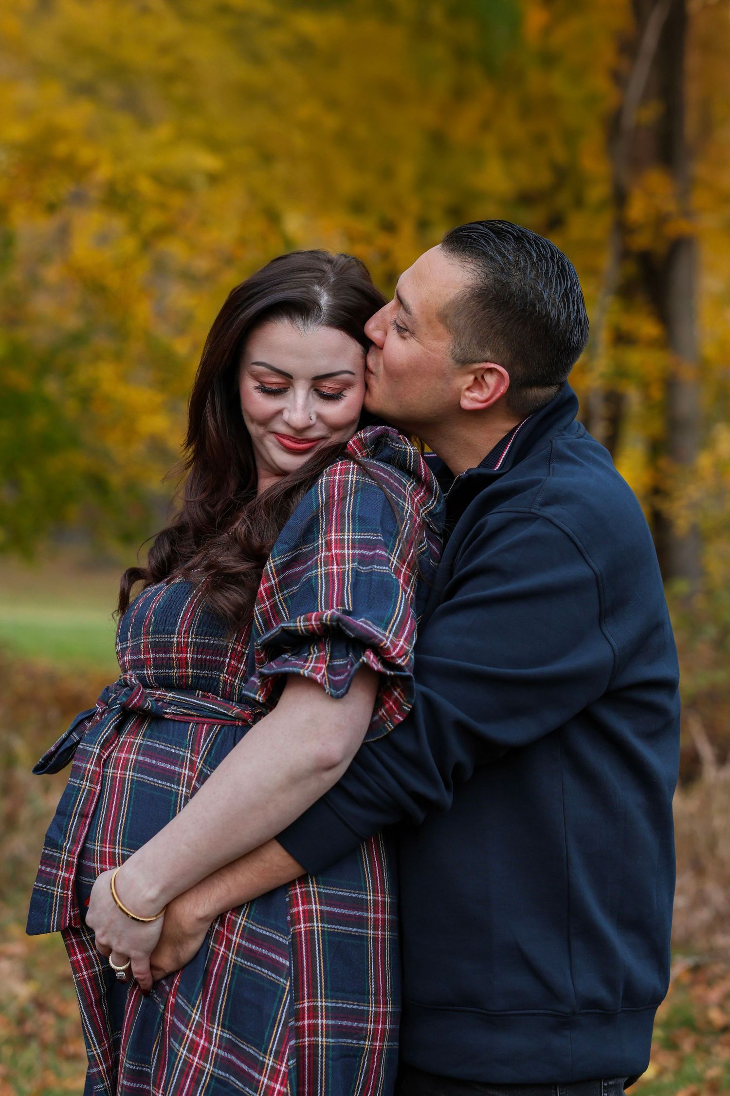 Man tenderly kisses pregnant woman's cheek while embracing her in fall park setting.