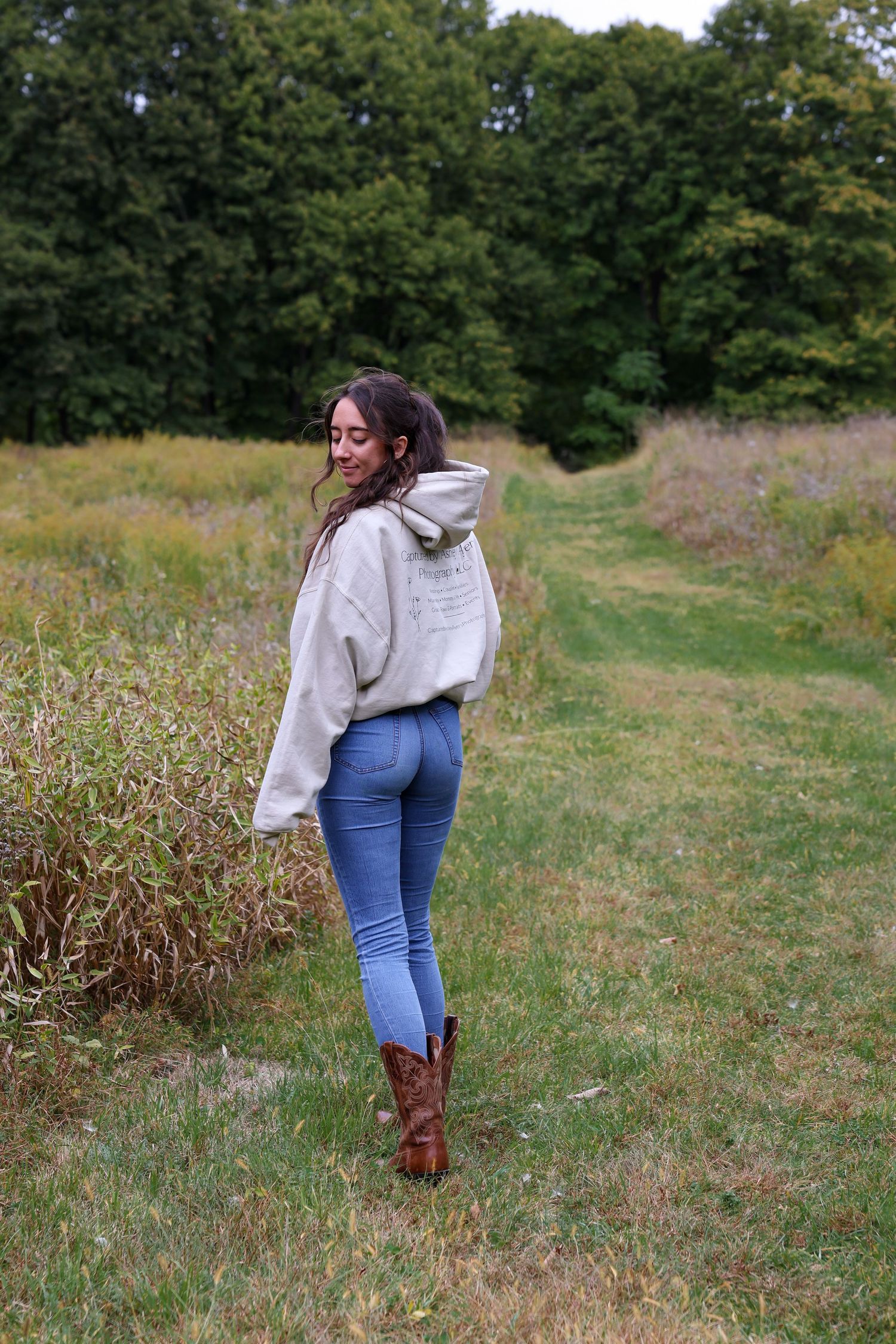 Young woman in gray hoodie, blue jeans, and brown cowboy boots standing in a green meadow with trees.