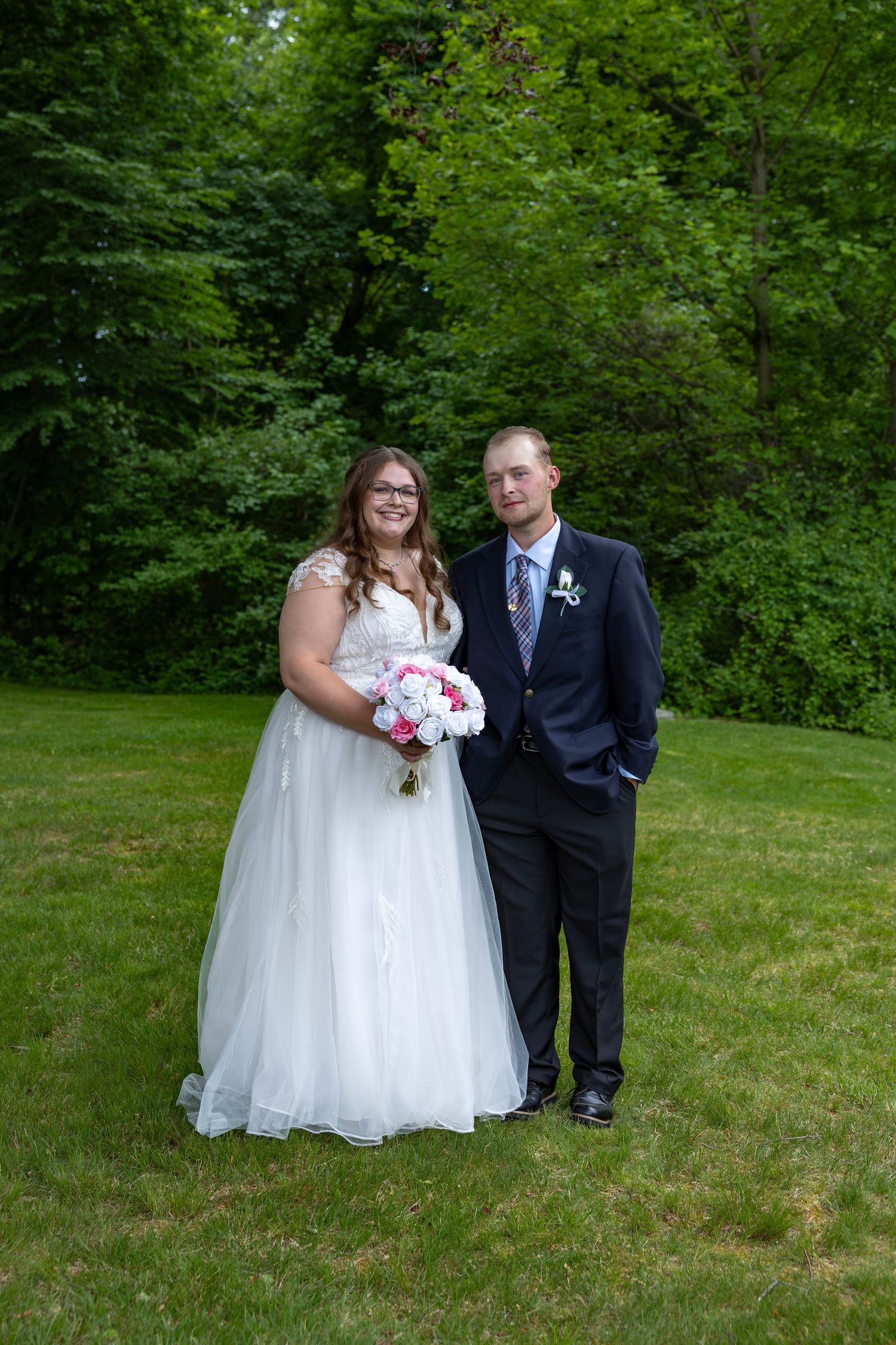 Bride in white lace gown holding pink bouquet stands with groom in navy suit on green lawn.