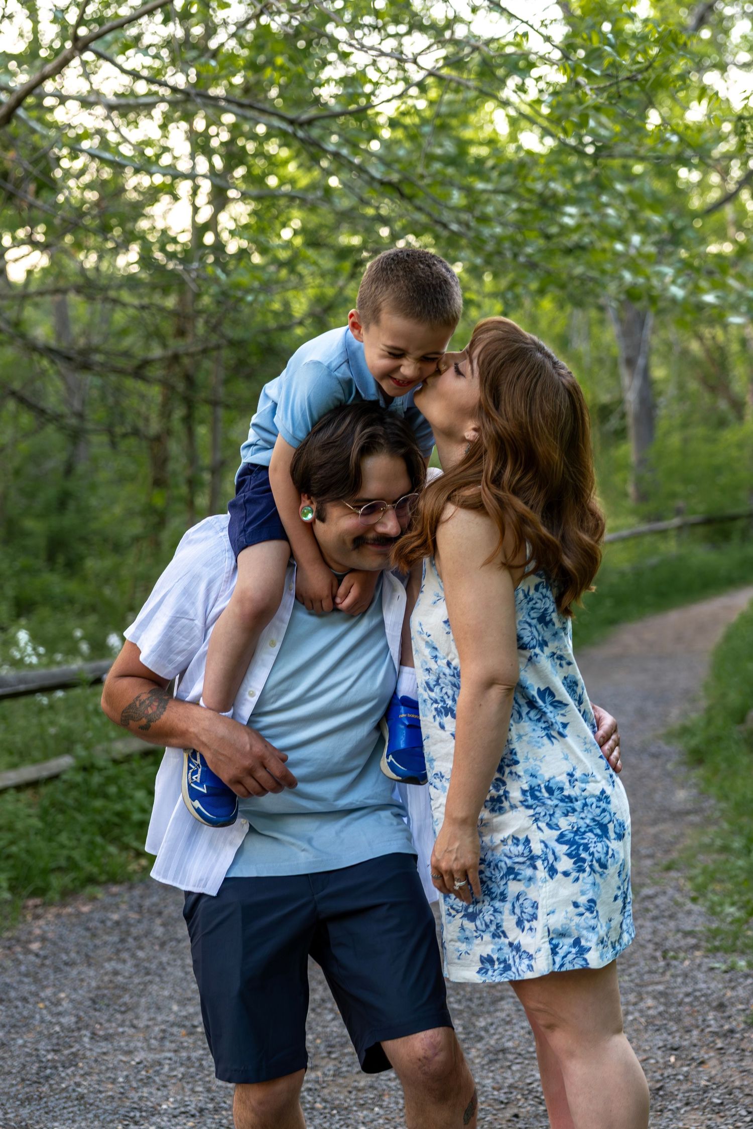 Happy family moment as young boy sits on fathers shoulders while mother kisses childs cheek on a park path.