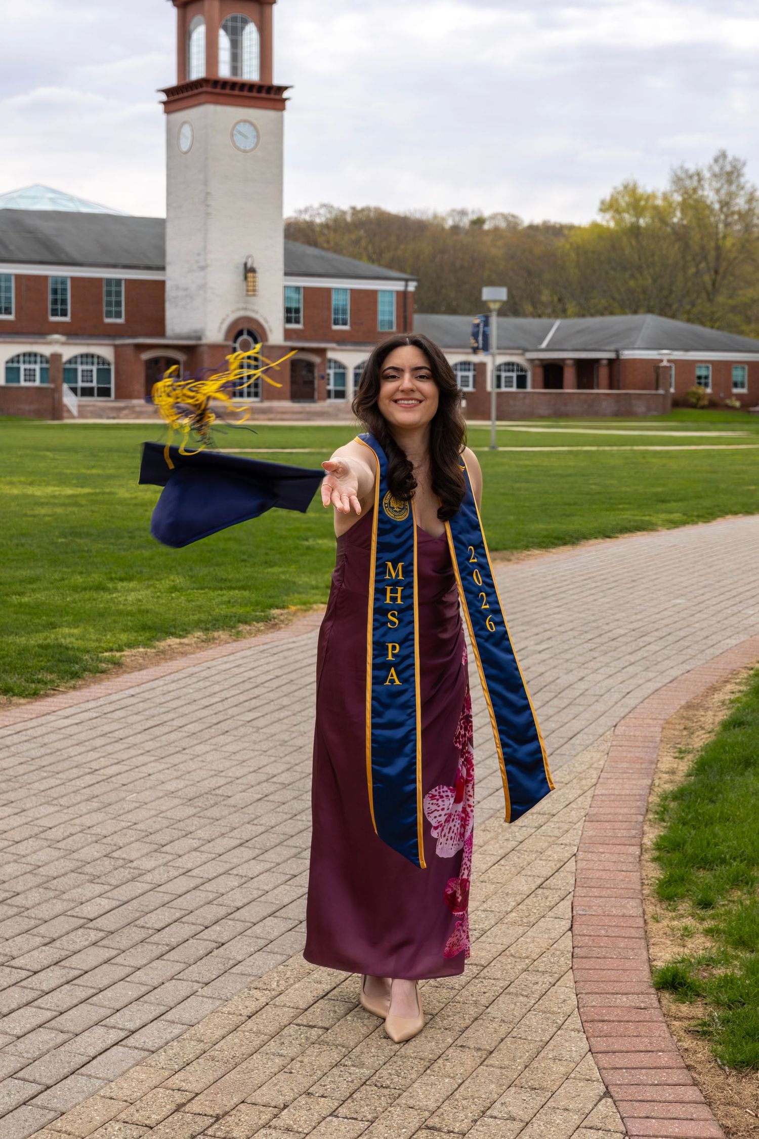 Female graduate in burgundy dress with blue sash tossing cap near campus clock tower.