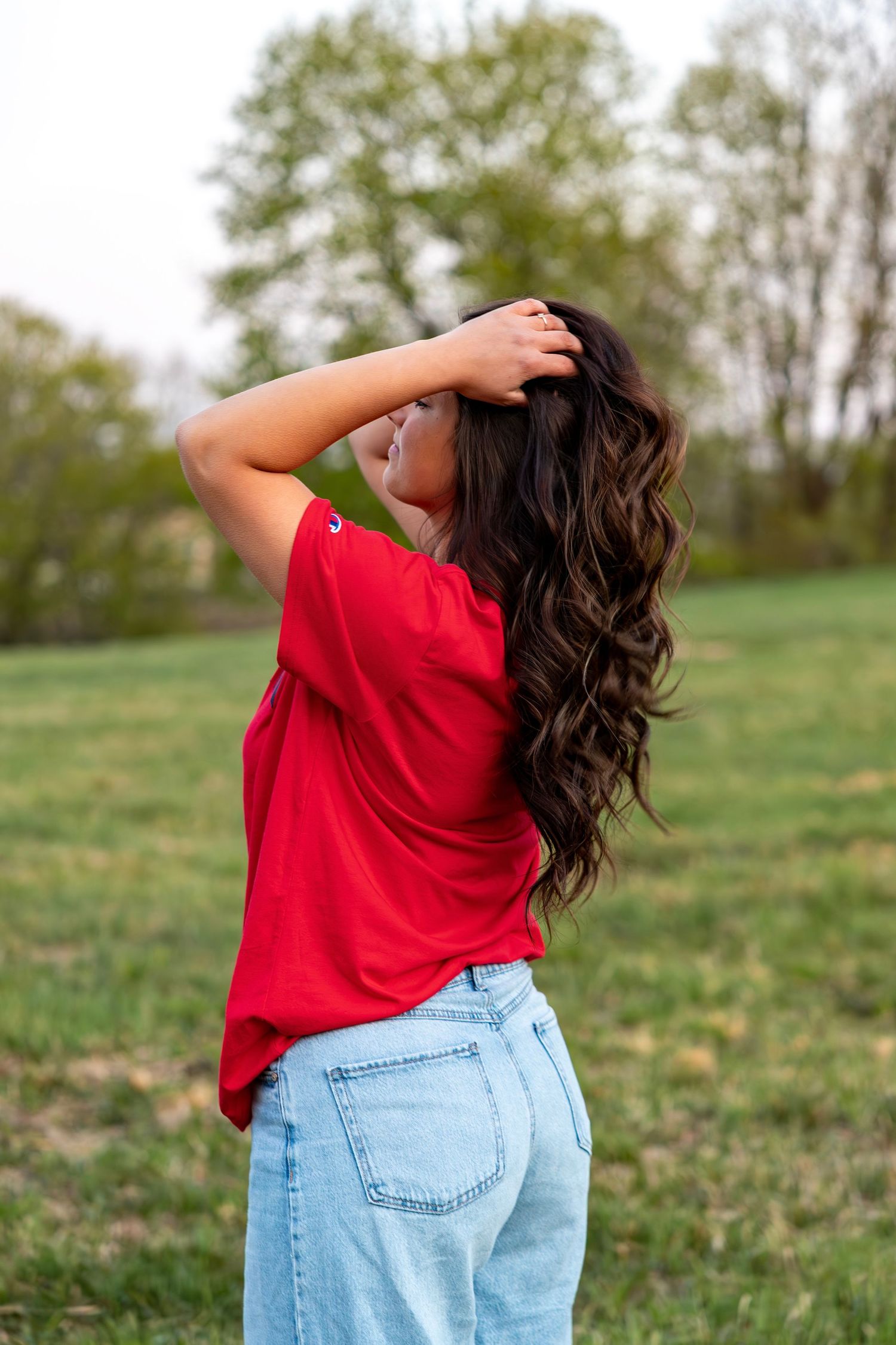 Back view of young woman with long curly dark hair in red tee and jeans in green outdoor field.