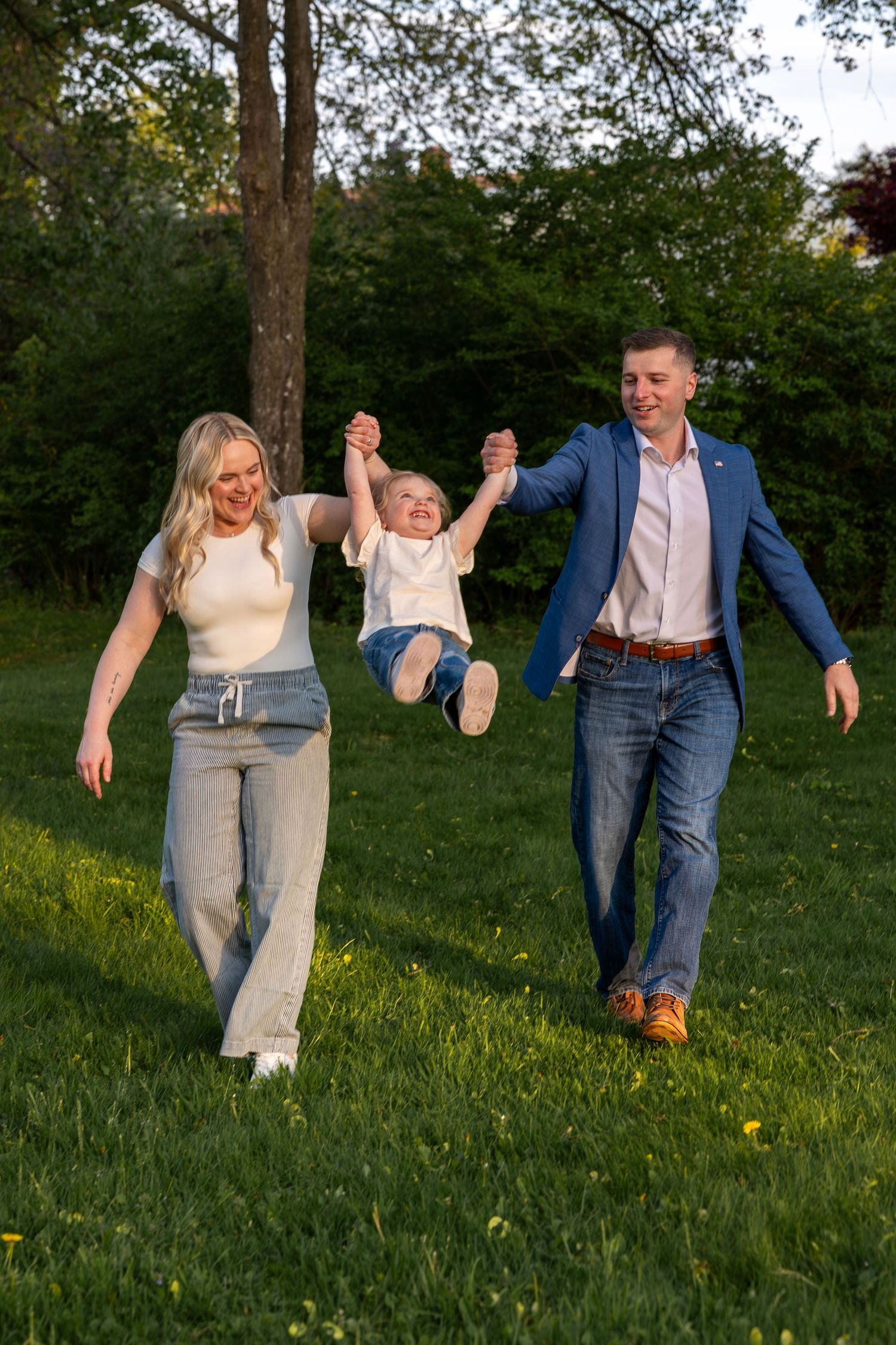 Joyful parents swinging laughing toddler between them while walking on sunny green lawn at golden hour.