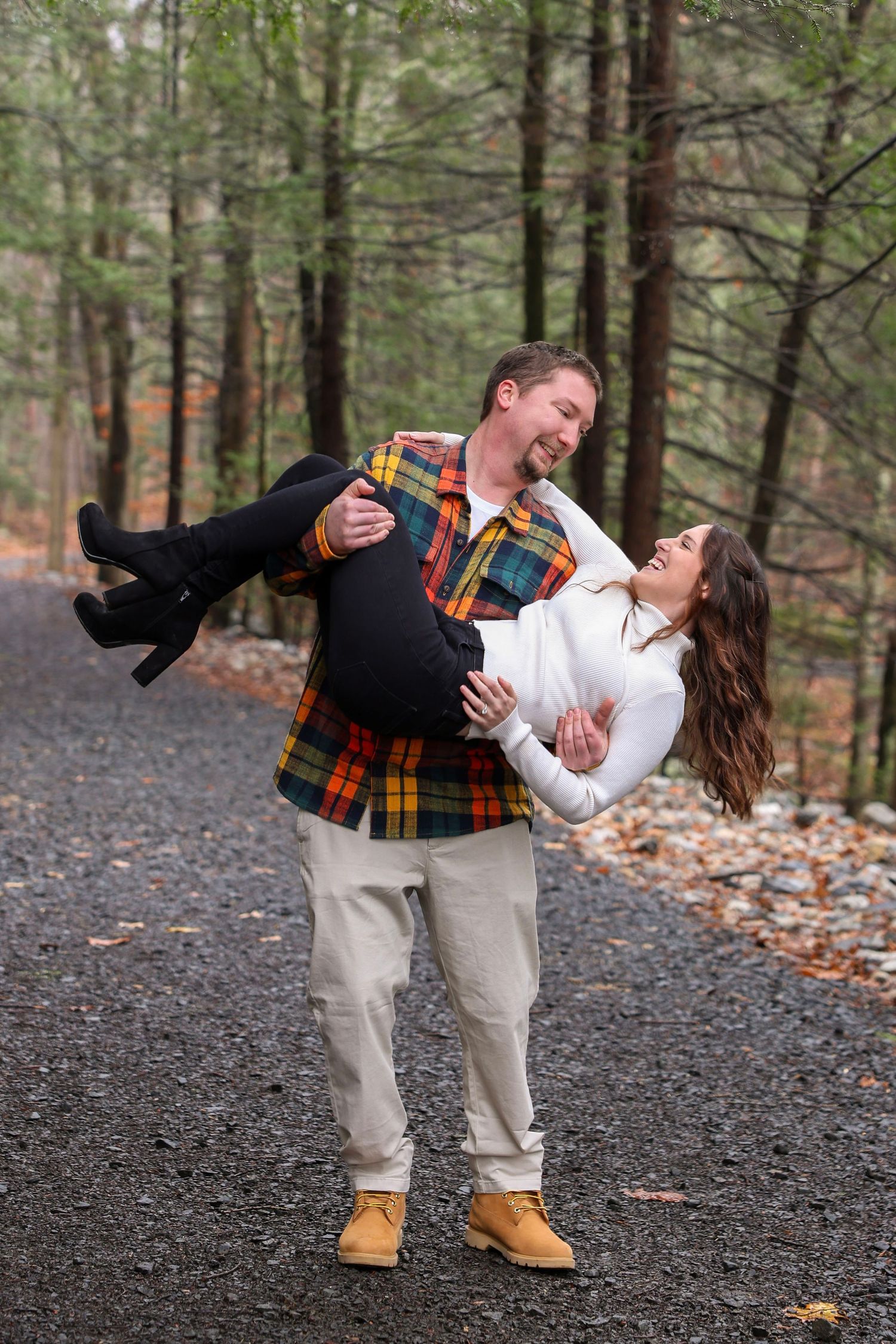 A man carries a laughing woman in his arms on a gravel path through a lush autumn forest setting.
