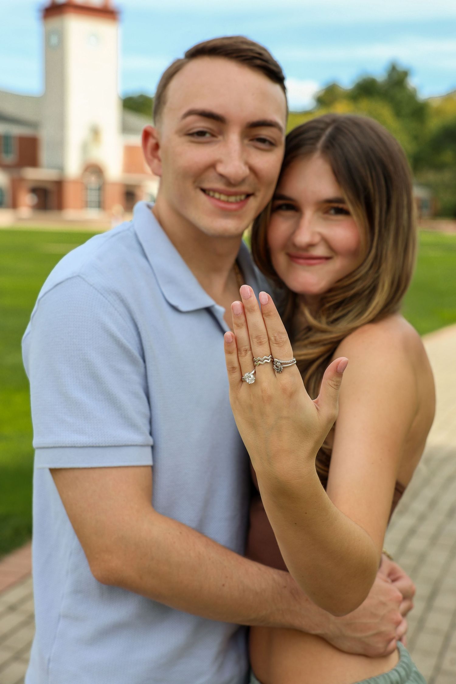 Happy engaged couple showing off engagement ring while embracing in front of a brick campus building.