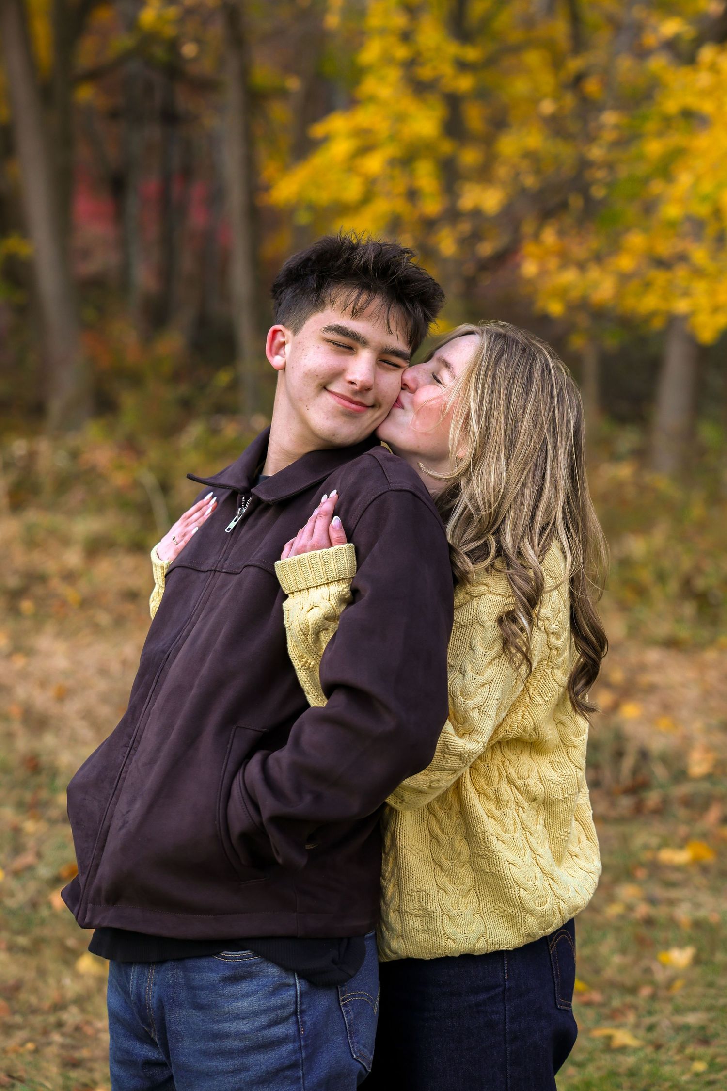 Young woman kissing mans cheek while embracing in colorful autumn park setting.