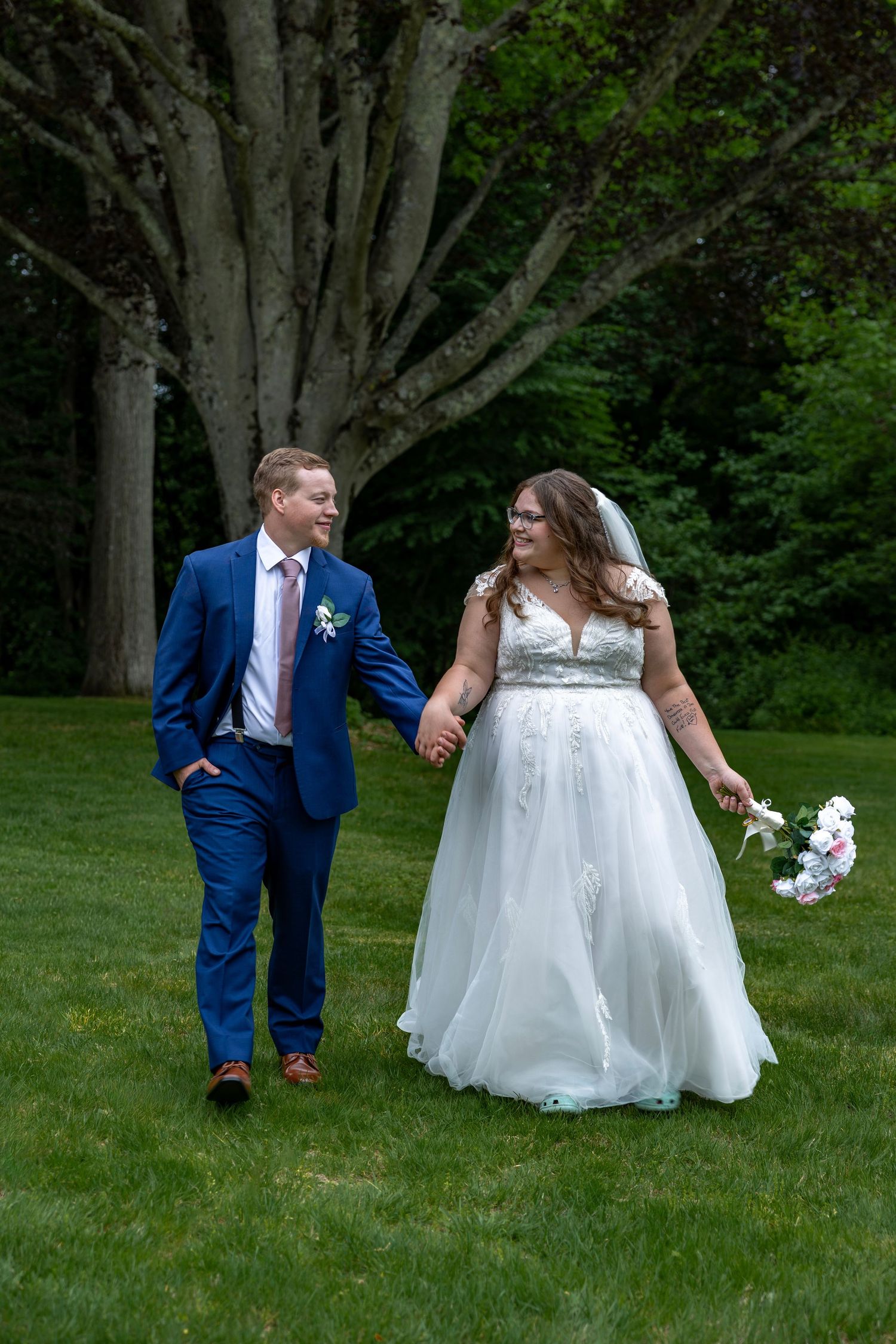 Bride and groom walk hand in hand on green lawn, smiling at each other in outdoor wedding portrait.