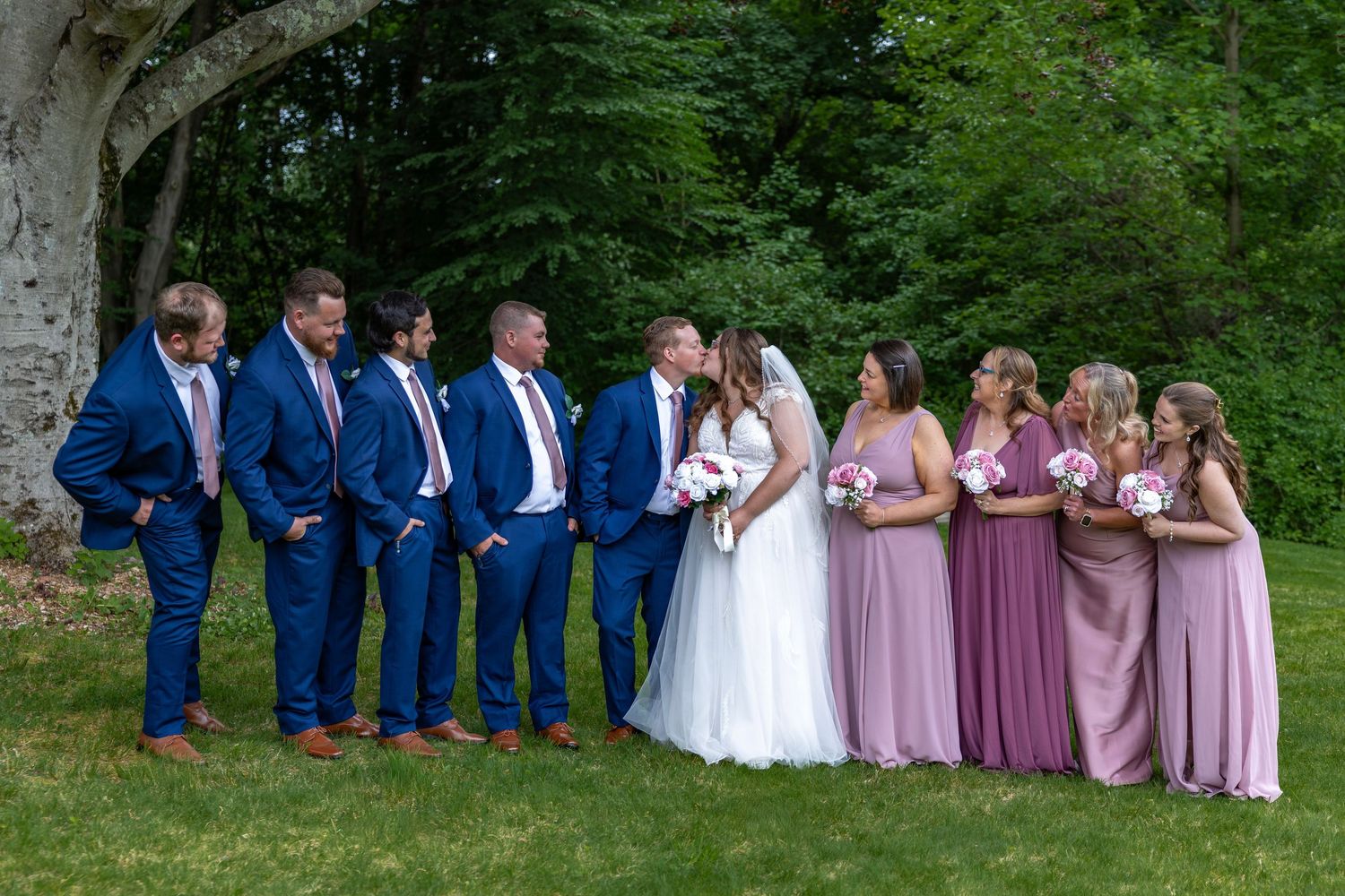 Wedding party poses outdoors with groomsmen in navy suits and bridesmaids in mauve.