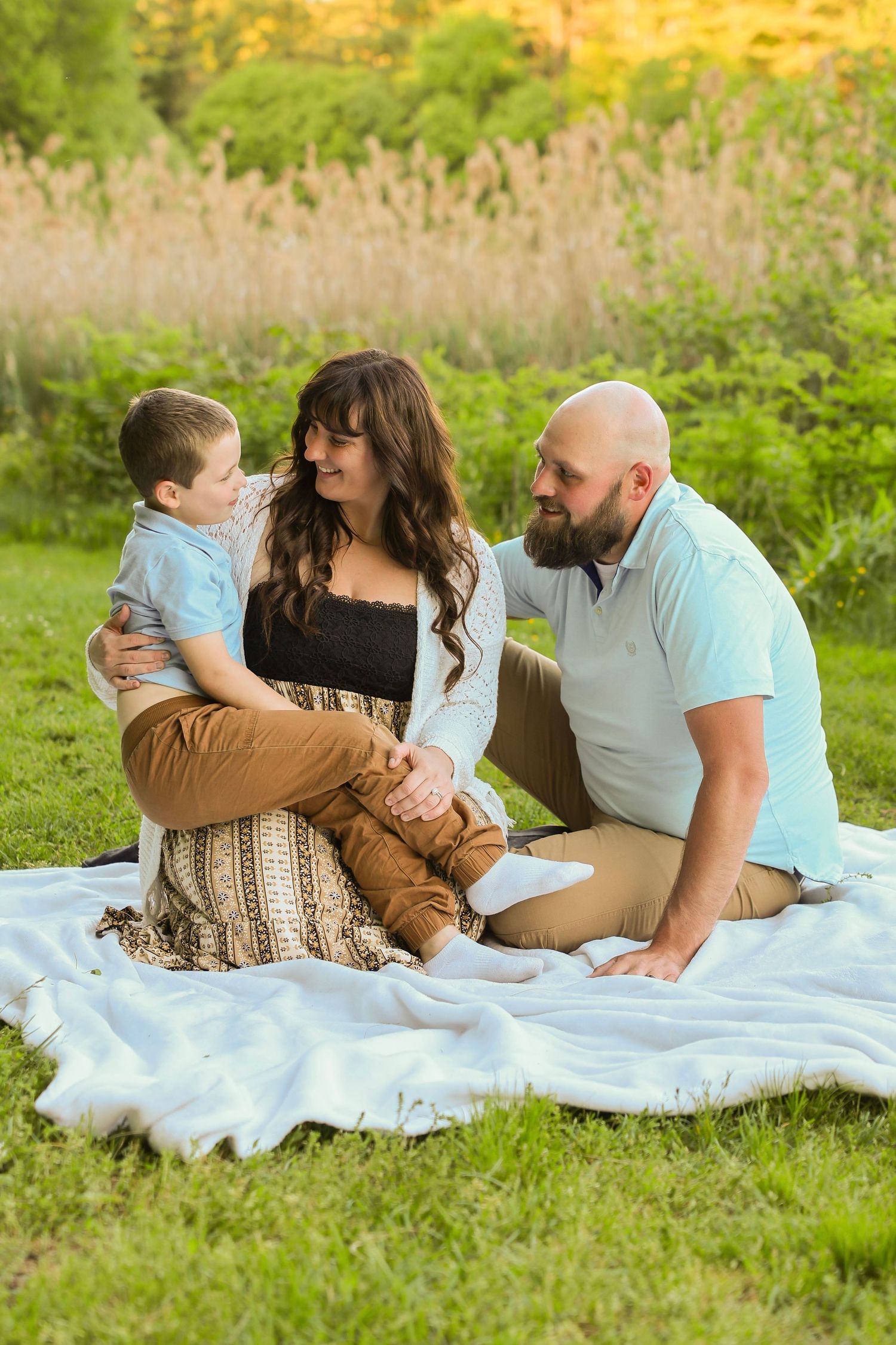 A loving family of three sits on a white blanket in a sunny meadow, sharing a warm moment.