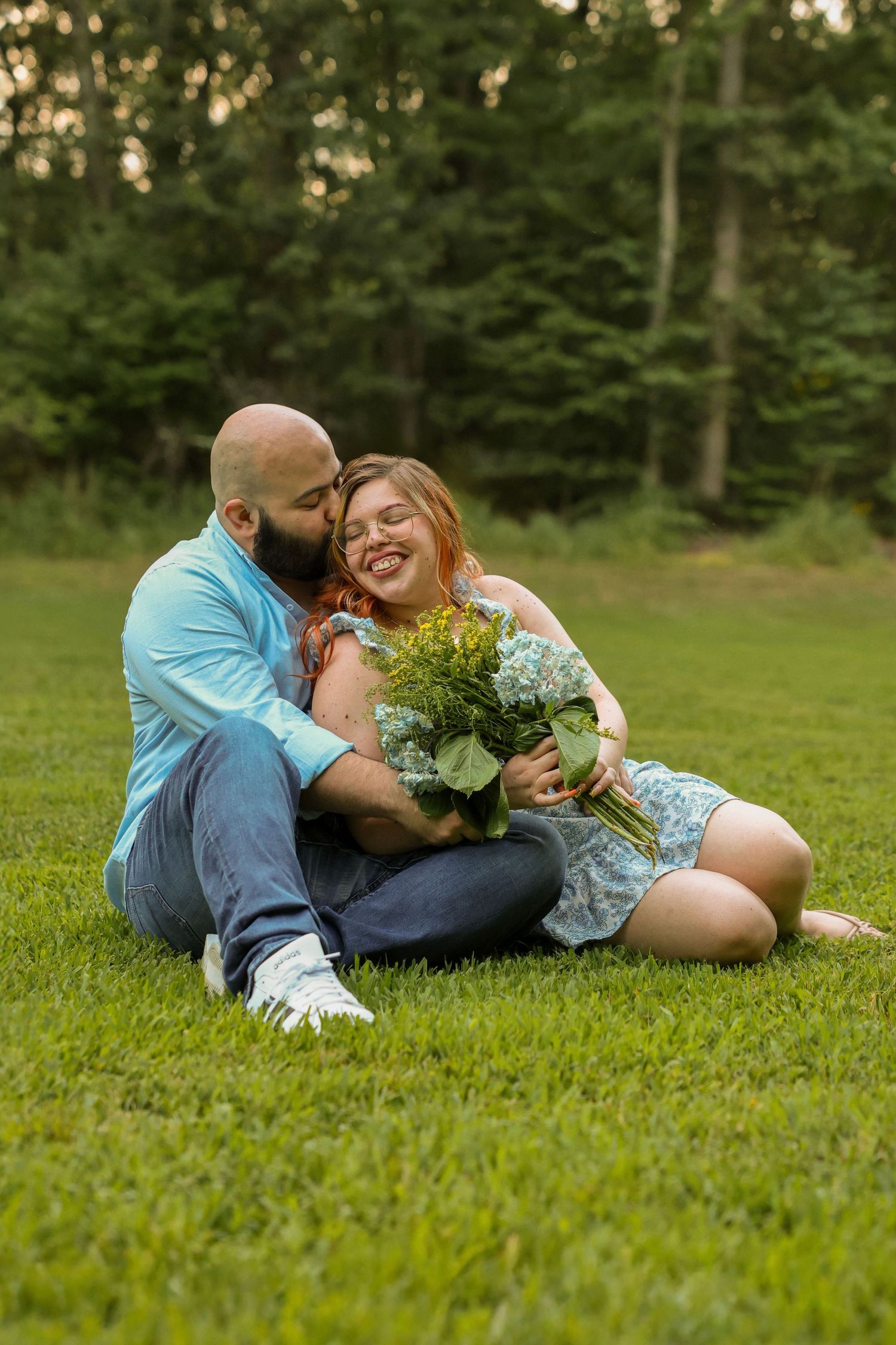 Happy couple sitting on grass, man kissing woman's cheek as she laughs holding a beautiful wildflower bouquet.