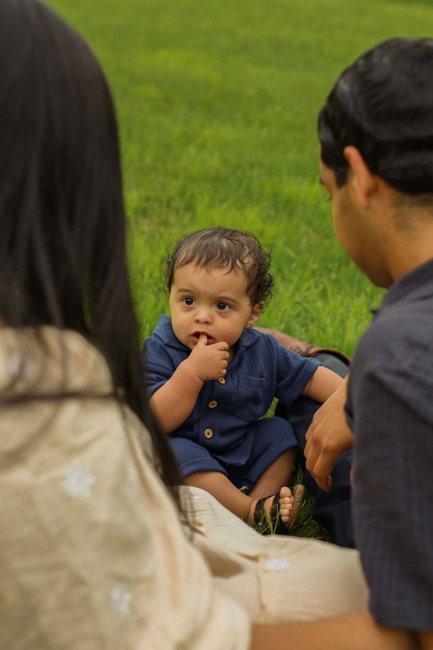 Baby with finger in mouth sits between parents on grass in outdoor portrait.