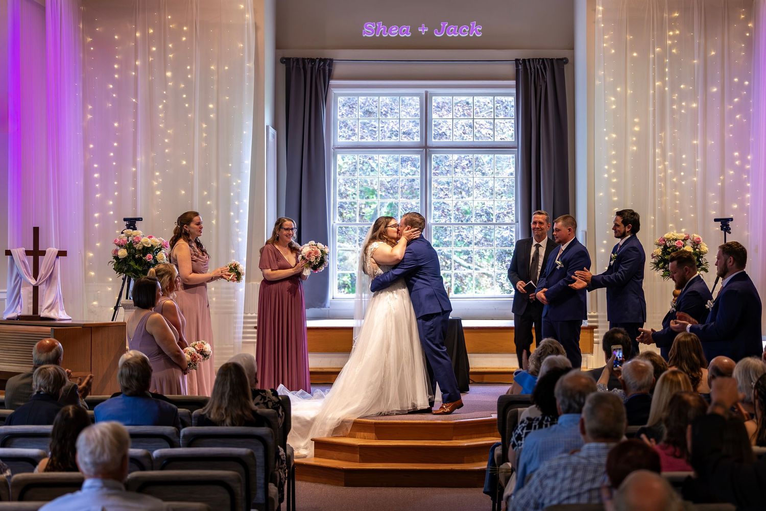 Bride and groom share first kiss at altar during wedding ceremony with bridesmaids and groomsmen in decorated chapel.
