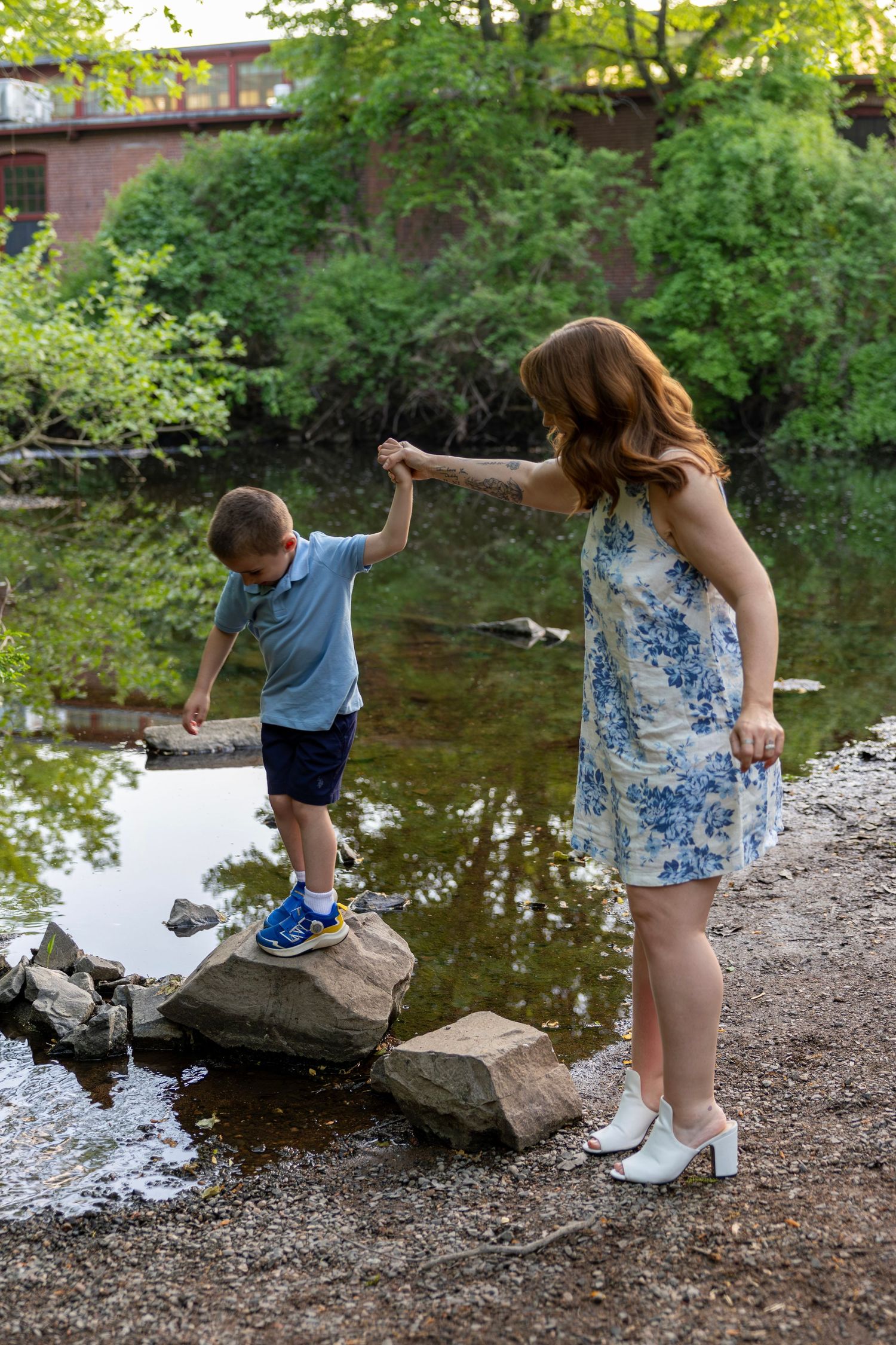 Mother holds sons hand as he carefully steps across rocks along a peaceful tree-lined riverbank.