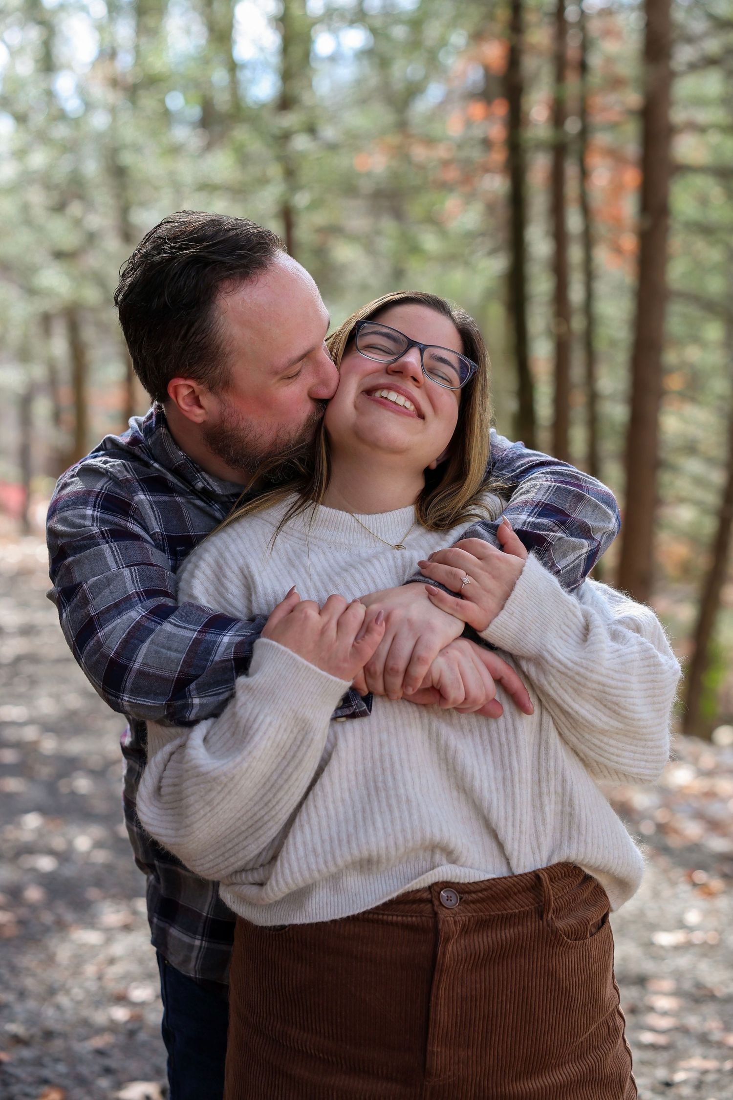 A couple embraces on a forest path, man kissing woman's cheek as she smiles joyfully in autumn woods.