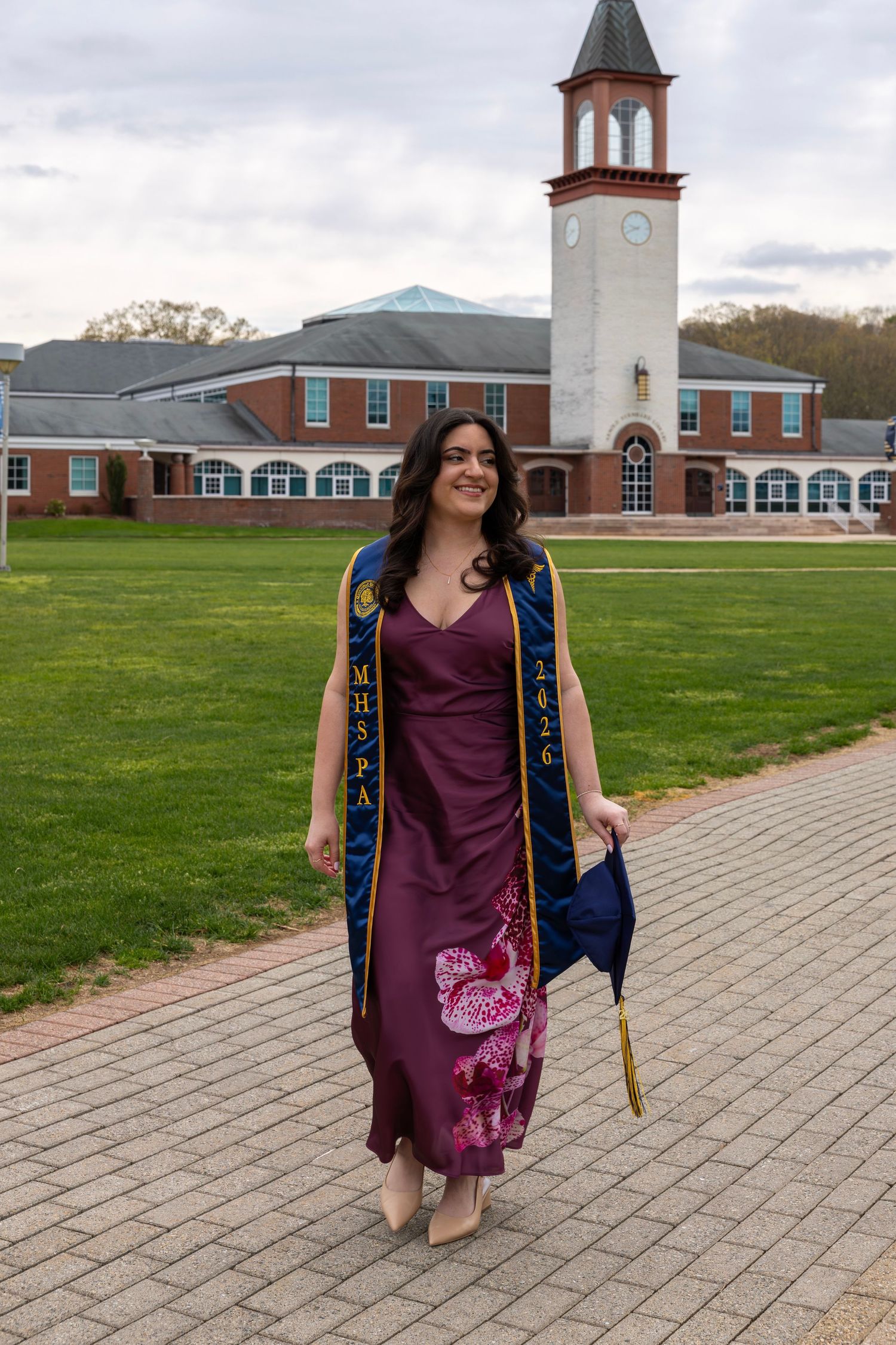 Female graduate in burgundy dress with blue MHSPA sash walking on brick campus pathway.