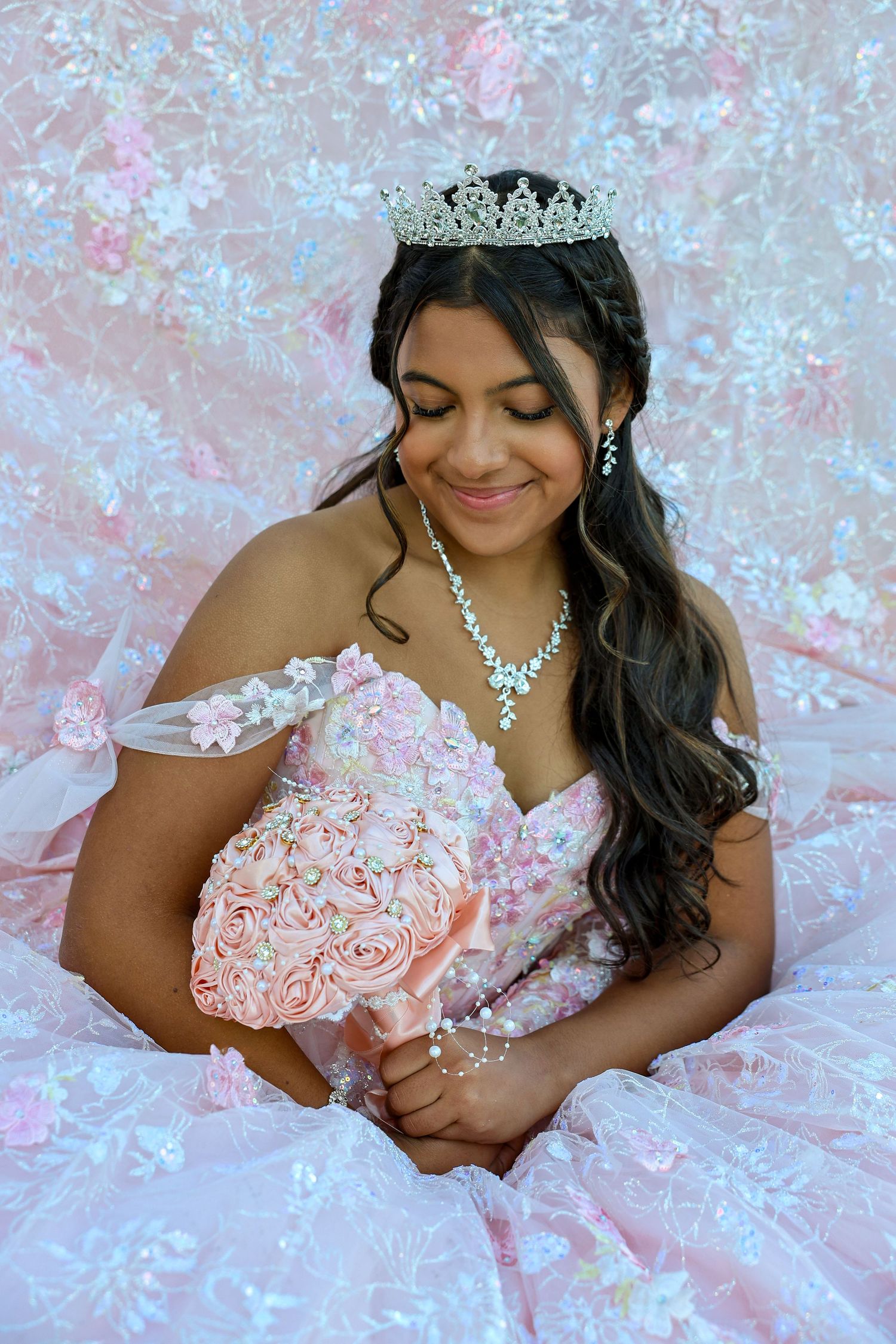 Smiling teenage girl in pink quinceañera ball gown holding floral bouquet wearing crystal tiara and necklace.