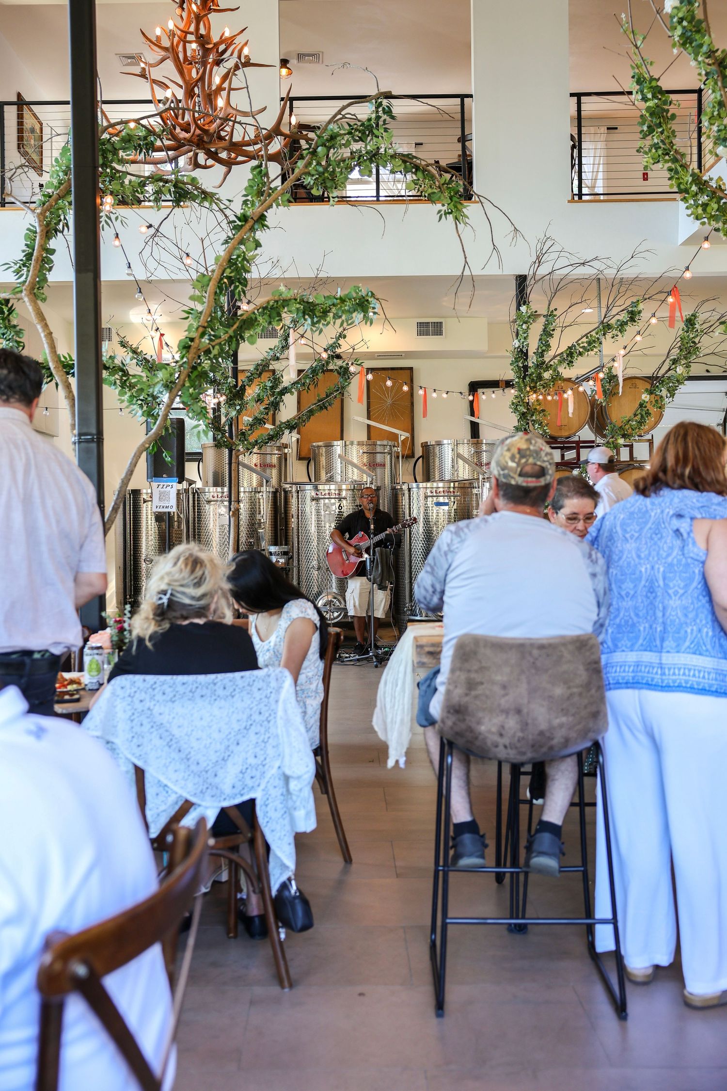 Guests dining inside a decorated brewery venue with greenery arches and a live musician performing.