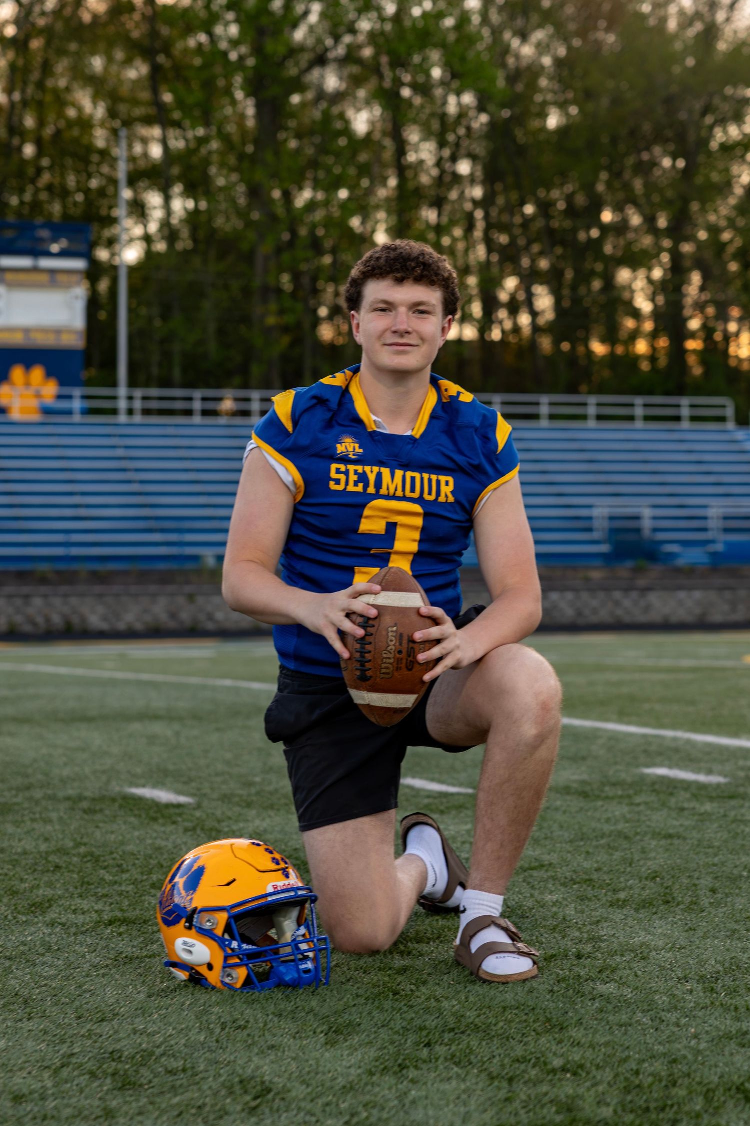 Seymour #3 football player kneeling on field holding football with blue helmet beside him at sunset.